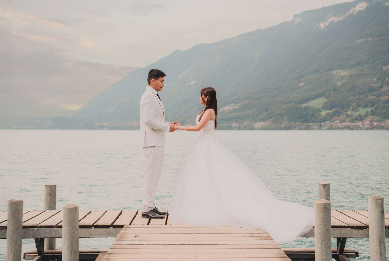 Bride and groom holding hands on a wooden dock with turquoise alpine lake and mountains backdrop.