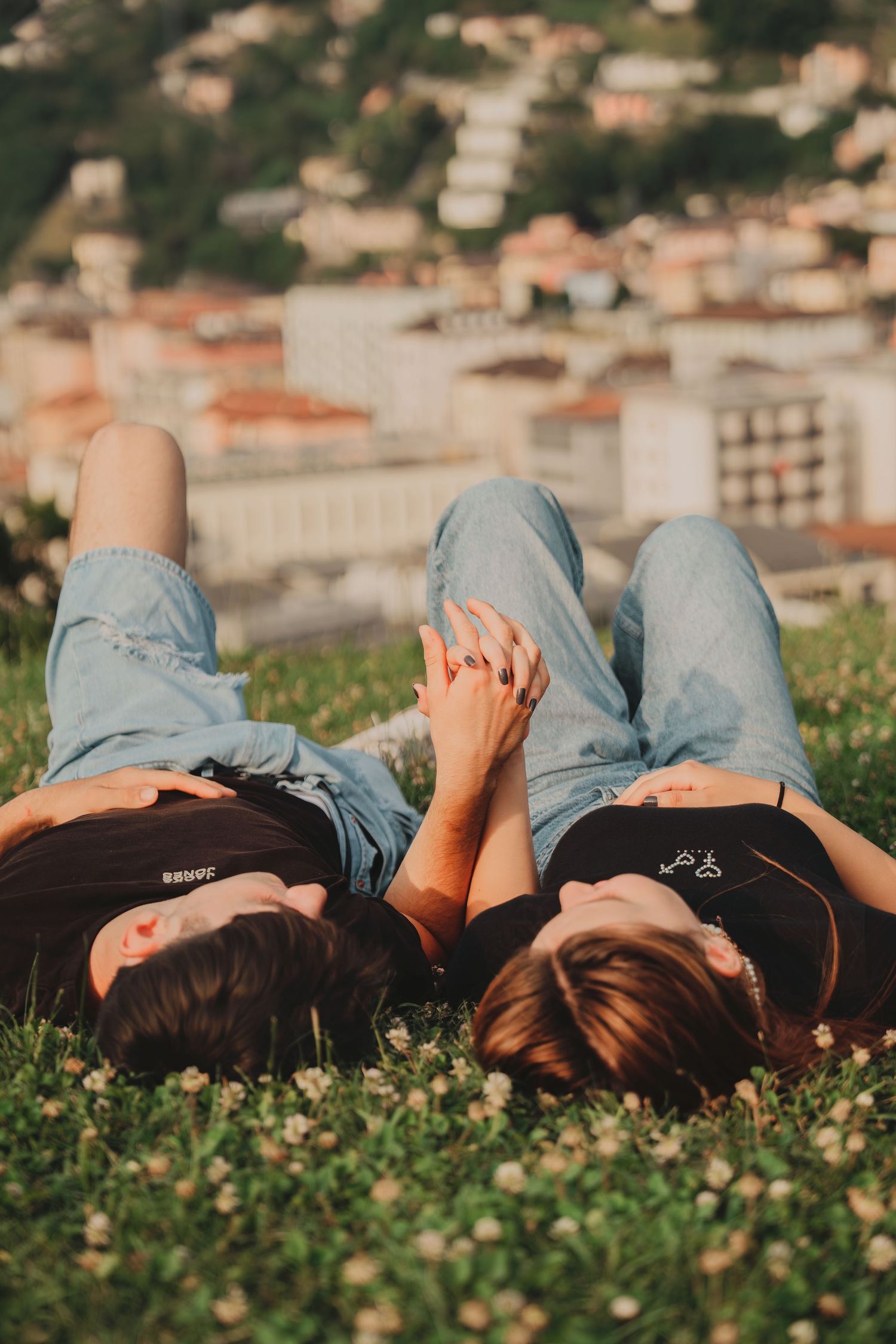 Couple lying on grass holding hands with city buildings visible in background.