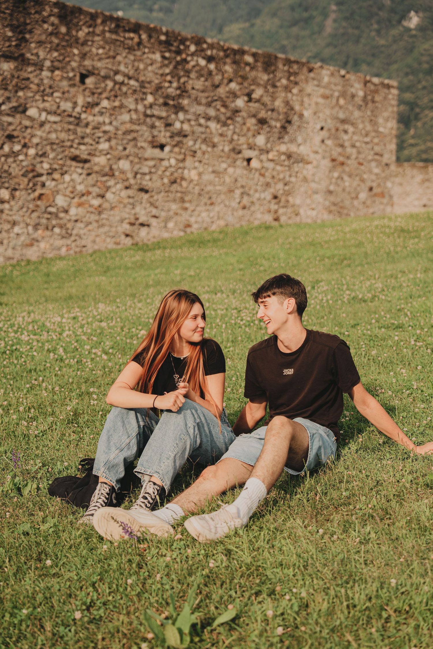 A smiling young couple sitting together on a sunny green lawn beside an ancient stone castle wall.