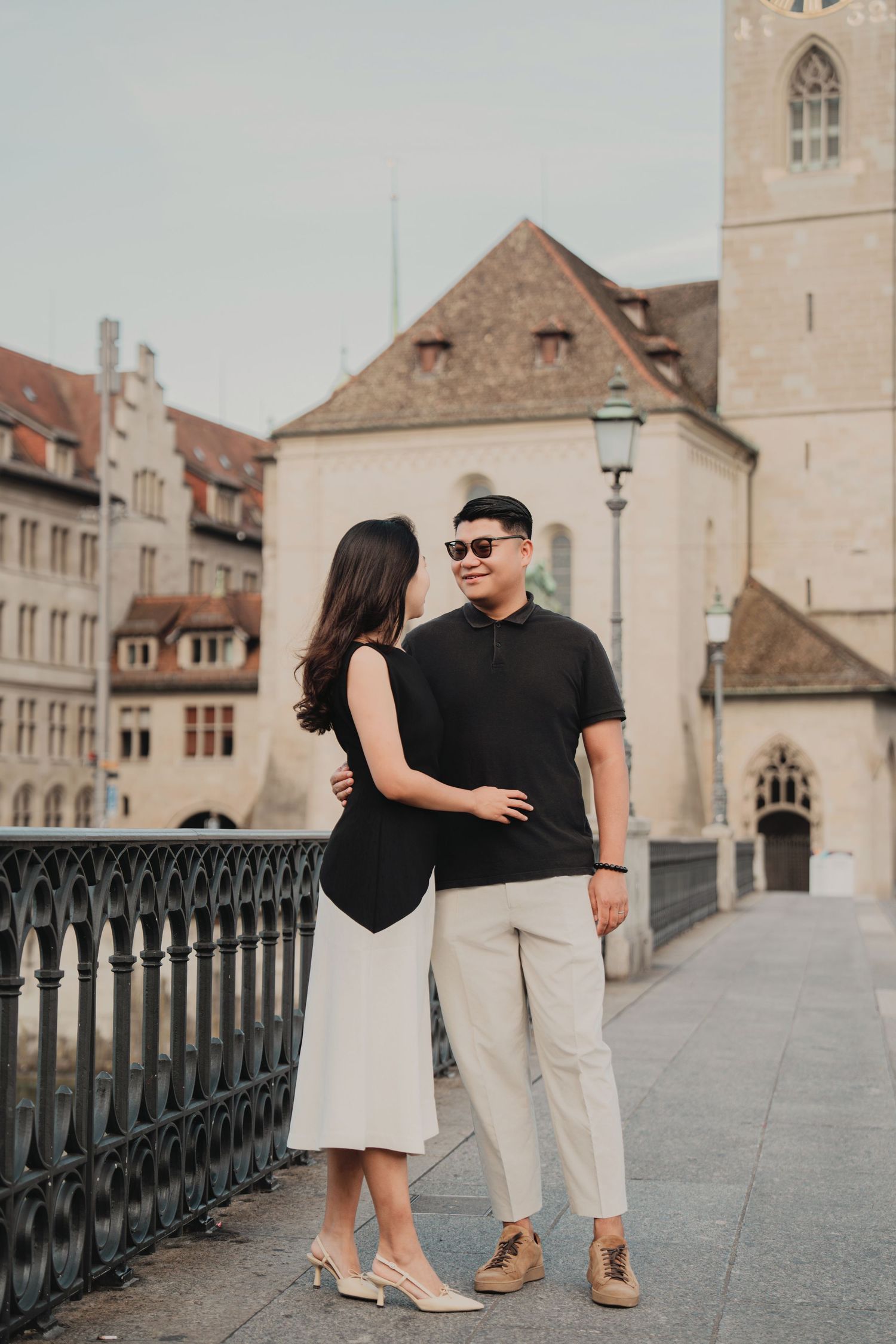 Couple embracing on a Zurich bridge wearing black tops, with a Gothic church and lamp post behind.