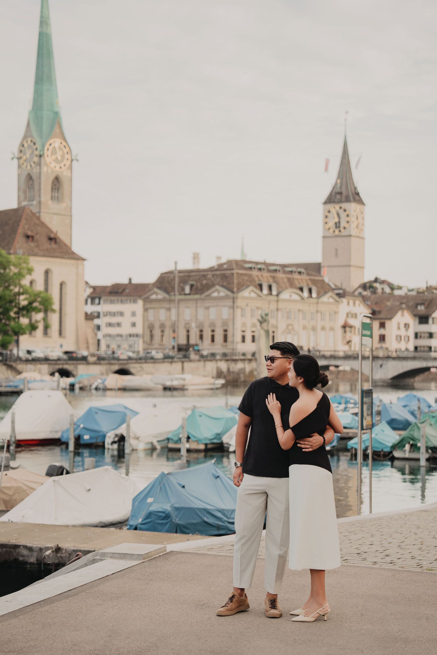 Couple embracing near Zurich harbor with Fraumunster and St. Peter's church towers in background.