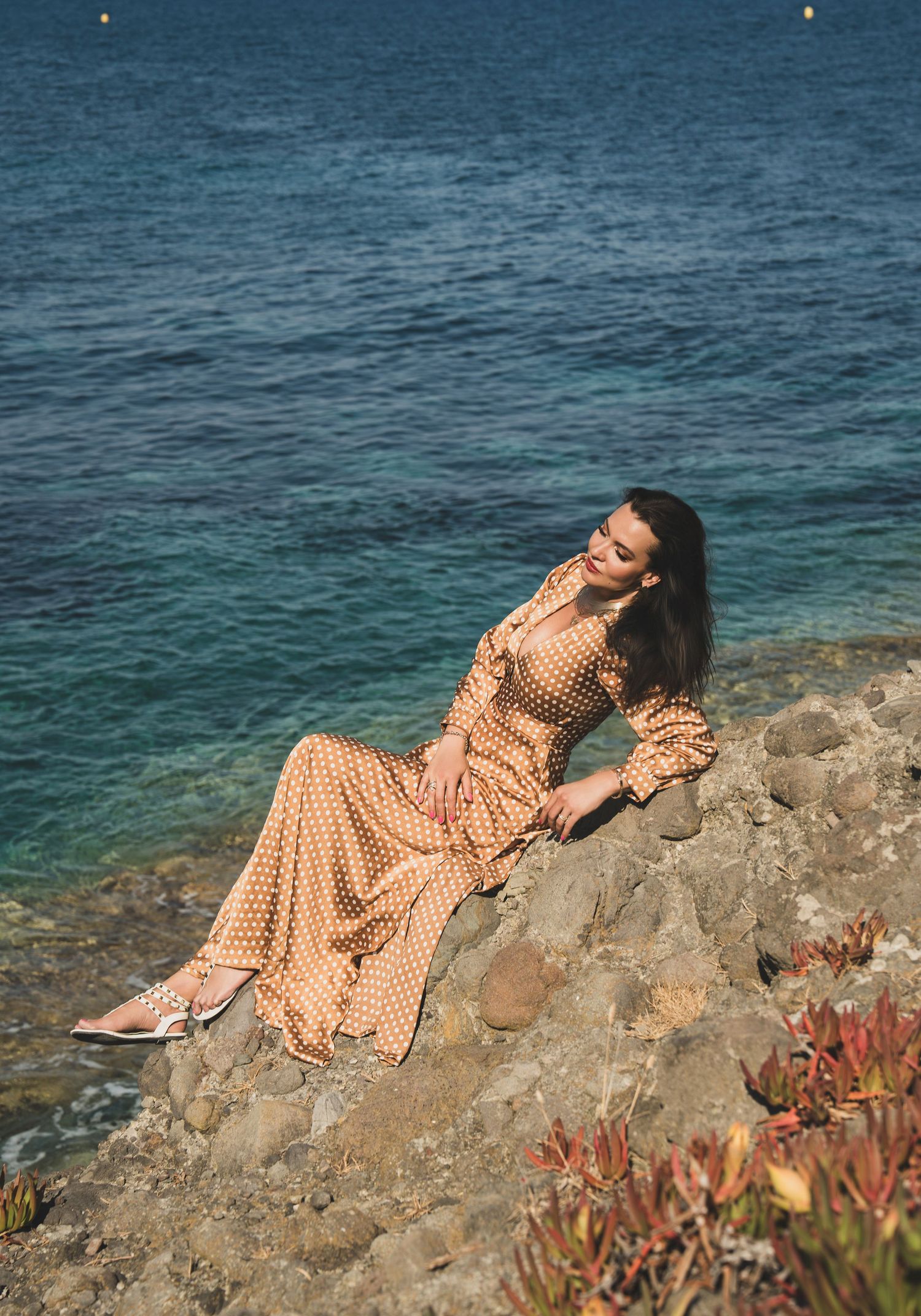 Woman in gold polka dot maxi dress reclines gracefully on sun-warmed coastal rocks by blue sea.