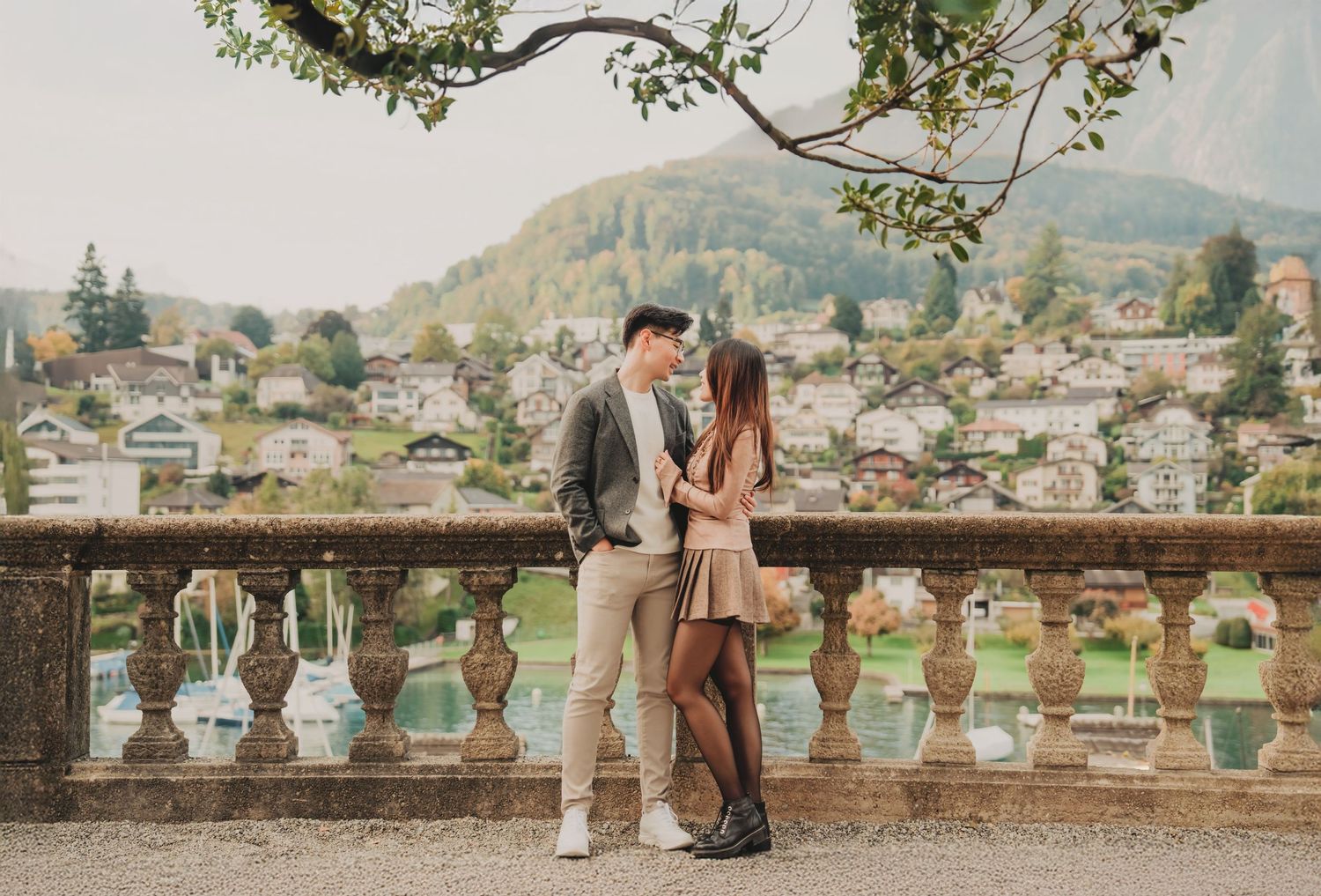 Couple standing close together on scenic stone terrace with Swiss village and lake backdrop.