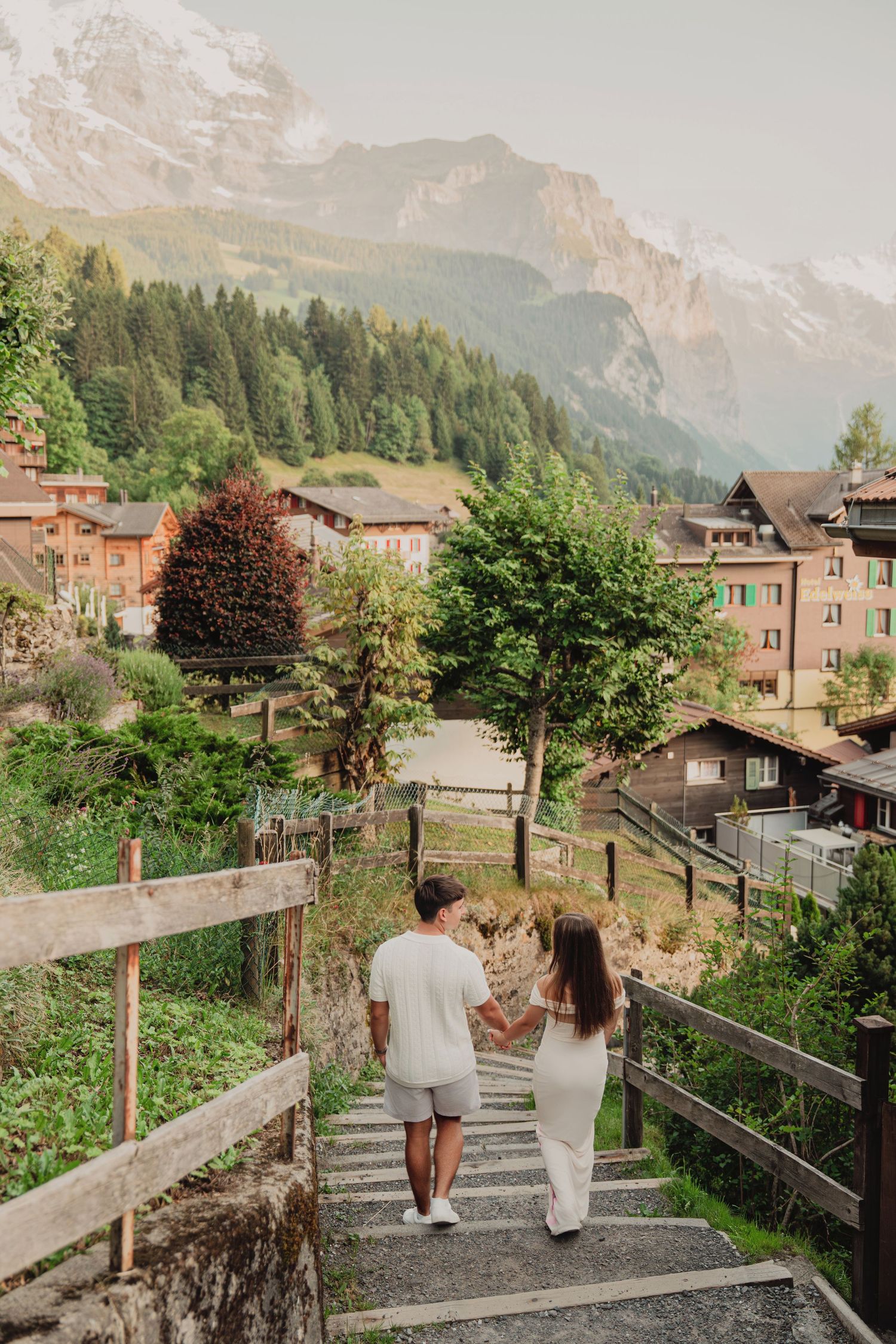 Young couple holding hands walking down stone steps in scenic Swiss mountain village with Alps backdrop.
