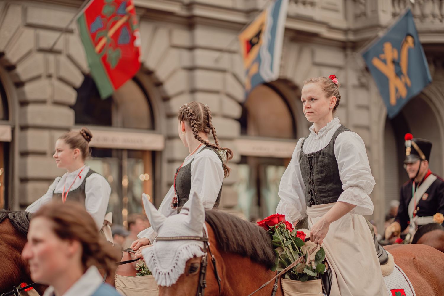 Young women in traditional Swiss dirndl dresses ride horses carrying roses in a parade.
