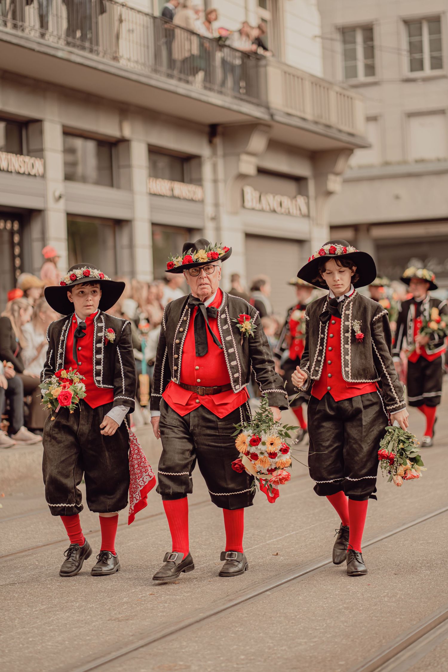 Three males in matching traditional Swiss black and red folk costumes march in a street parade.