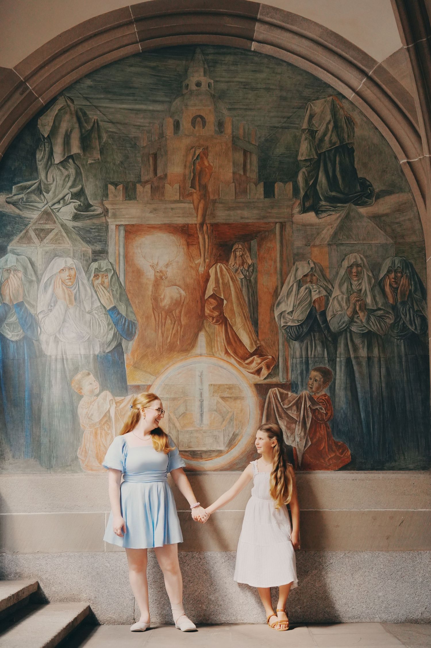 A mother and daughter in blue and white dresses hold hands in front of a large religious fresco mural.