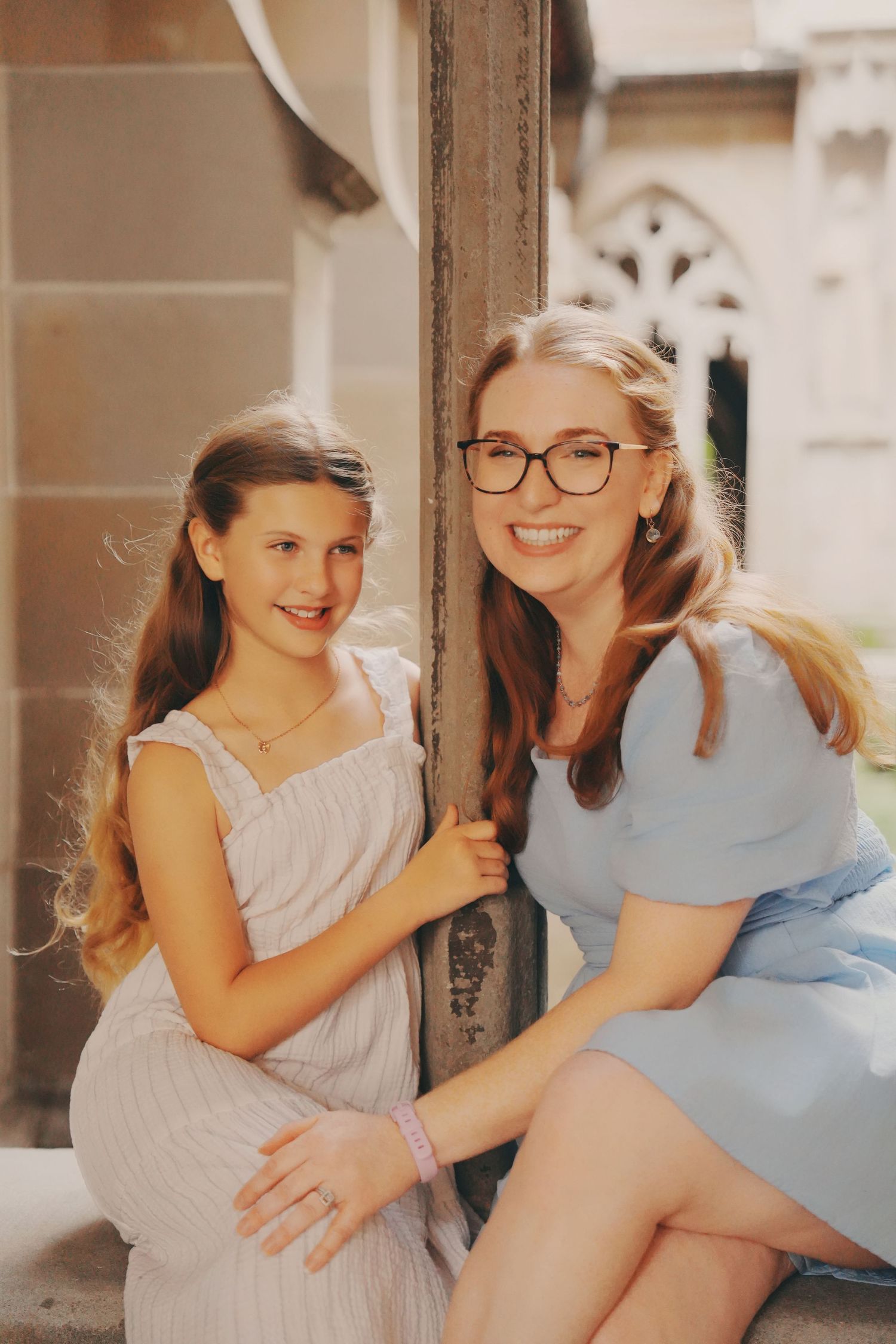 A smiling woman with glasses in a blue dress poses with a young girl in a white dress at a historic building.