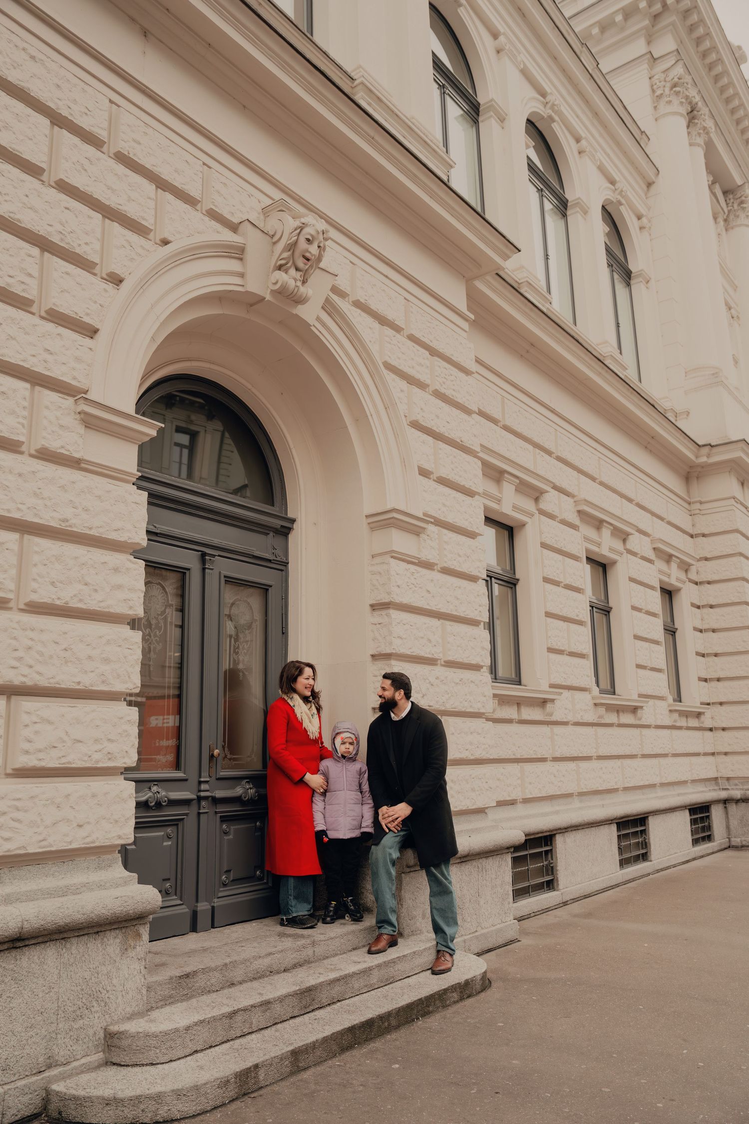 Family of three conversing on steps outside a classical European architectural building.