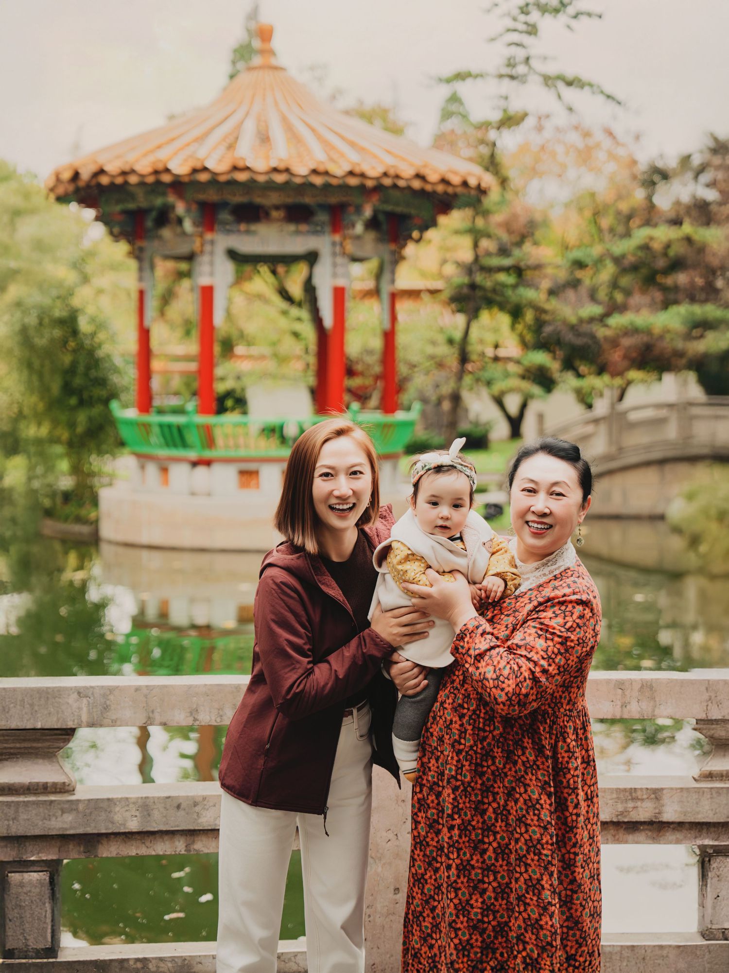 Three generations of women smiling with a baby near a Chinese garden pagoda.