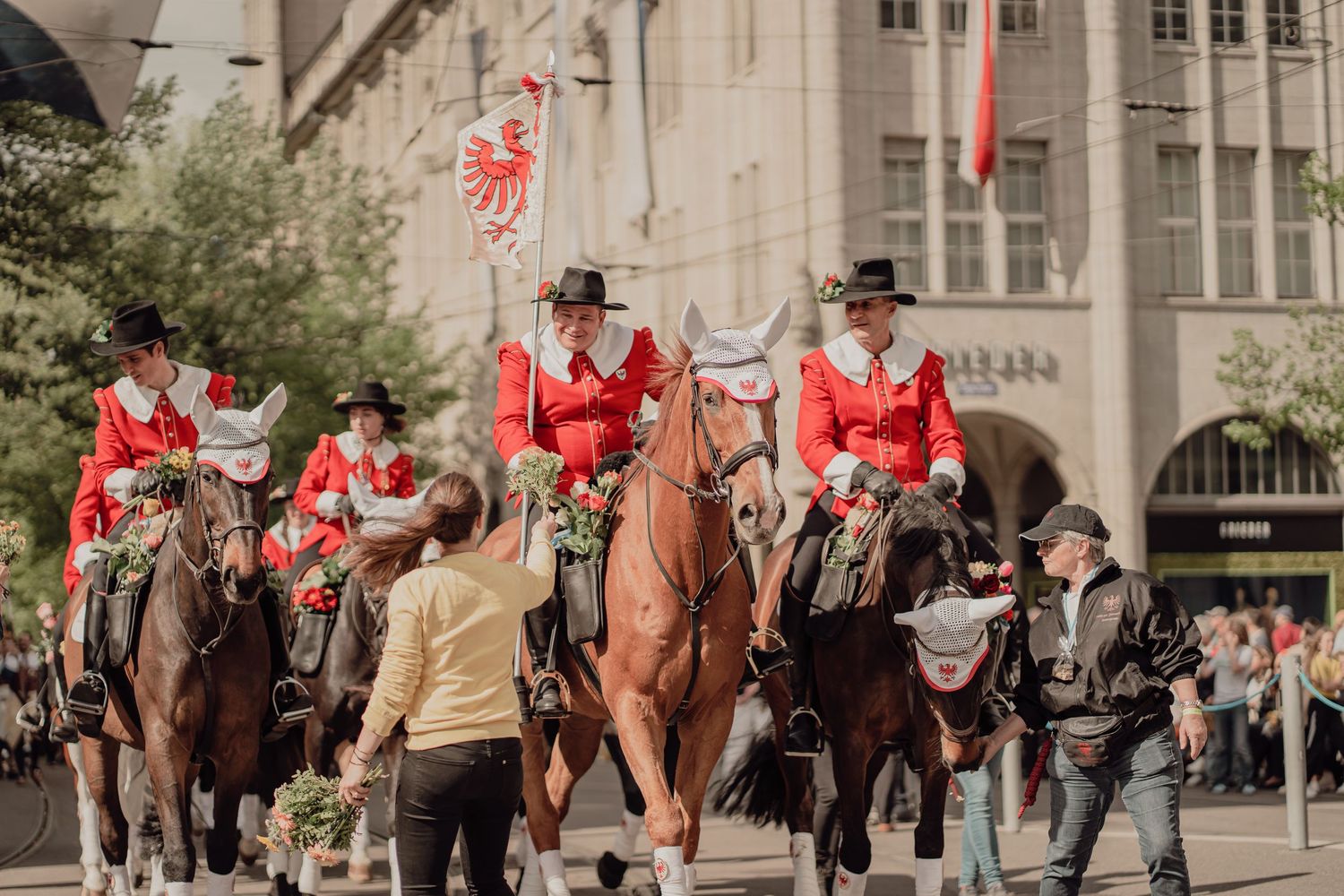 Equestrians in red traditional costumes ride decorated horses past spectators in a historic city parade.