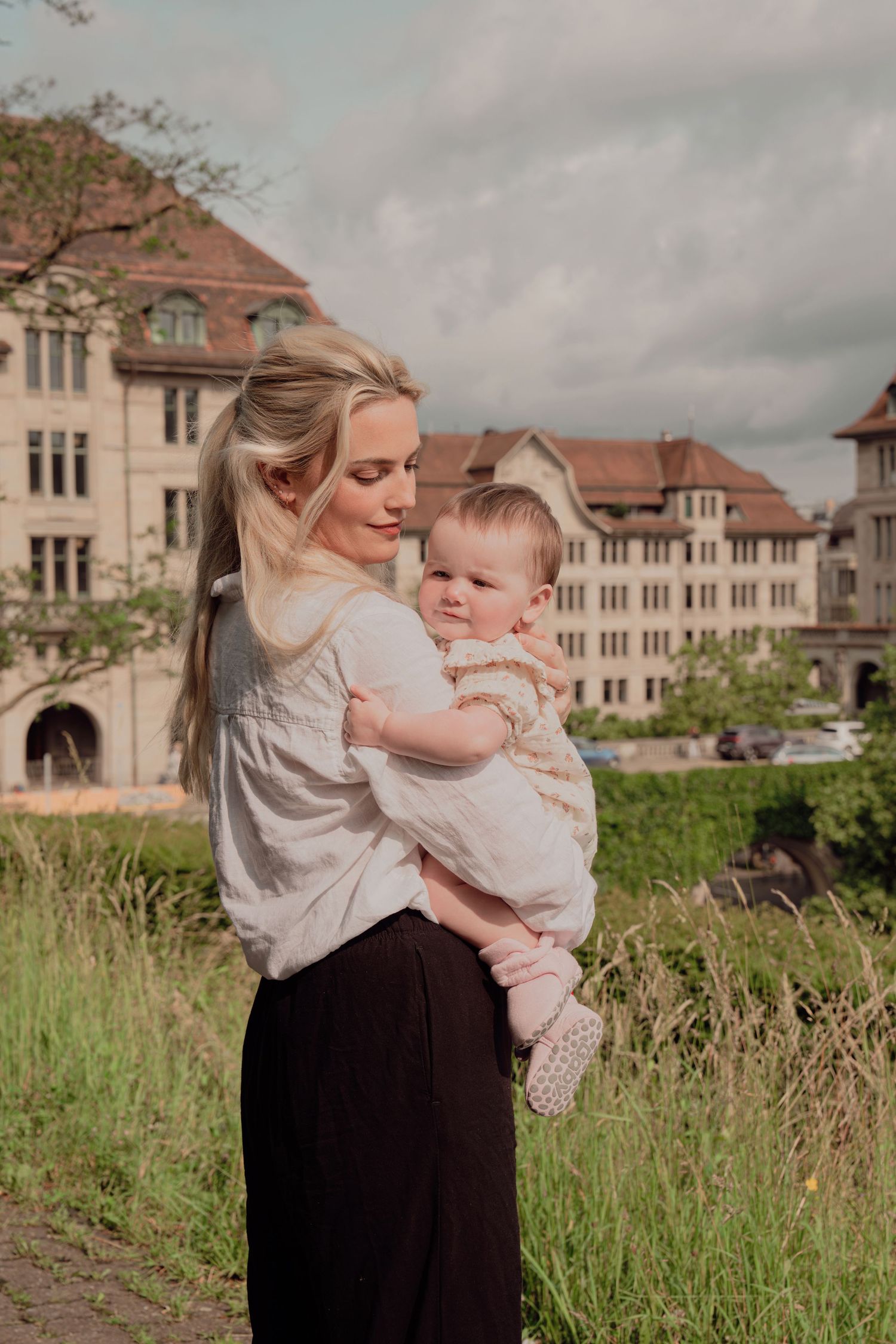 Tender moment as blonde mother gazes lovingly at baby she holds, European building in background.