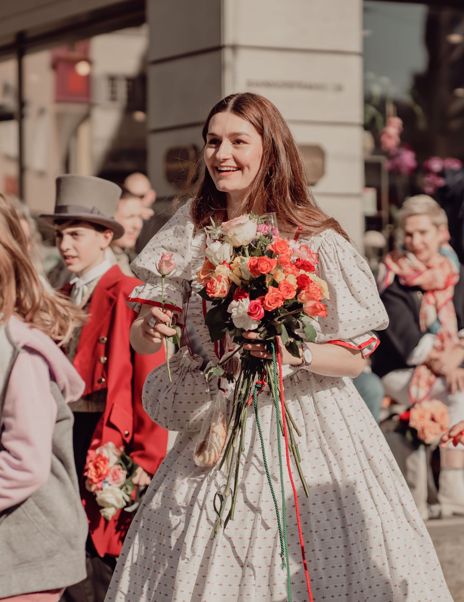 Smiling woman in a vintage floral dress holds a bouquet of orange roses during a street parade.
