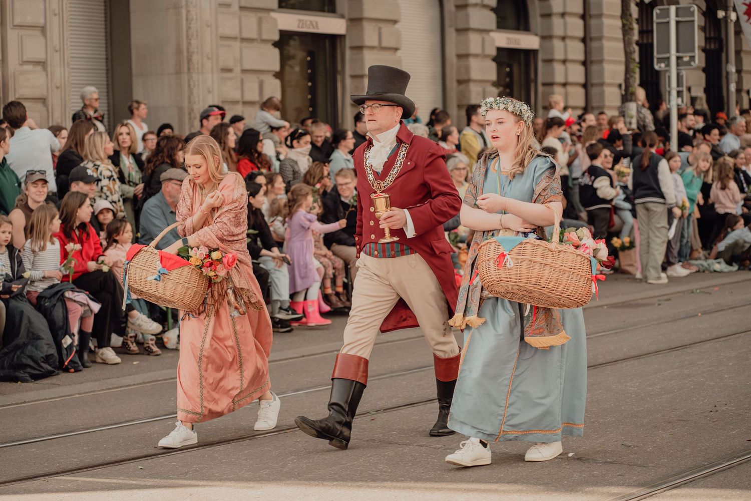 Man in top hat and red coat walks with two women carrying flower baskets in a historical street parade.
