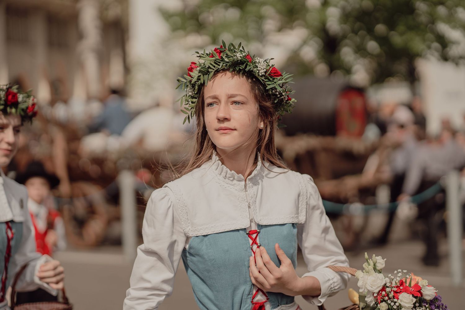 A young woman wearing a floral crown and traditional costume holds flowers during an outdoor historical parade.