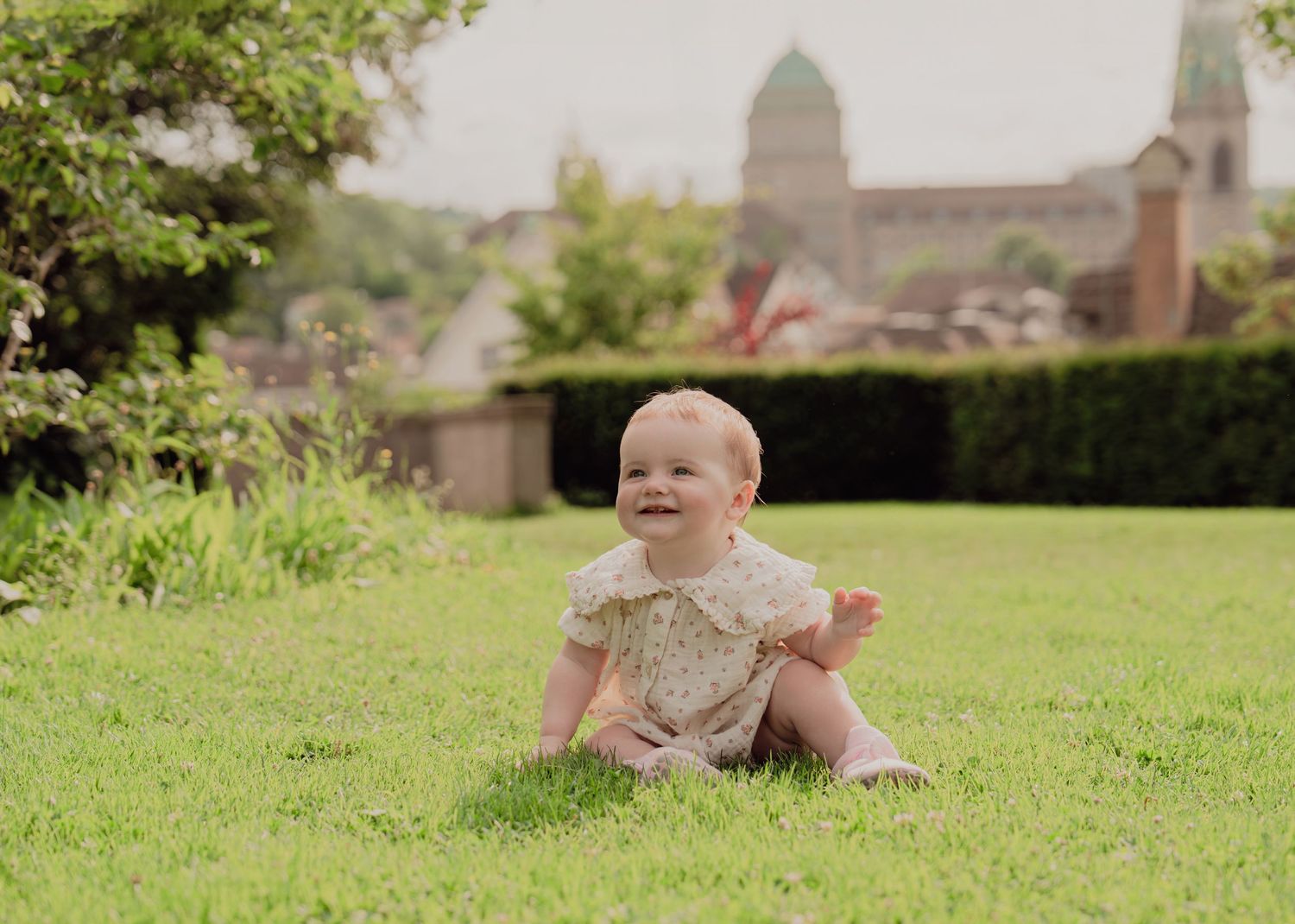 Cheerful baby in floral romper laughing on green lawn with European architecture behind.
