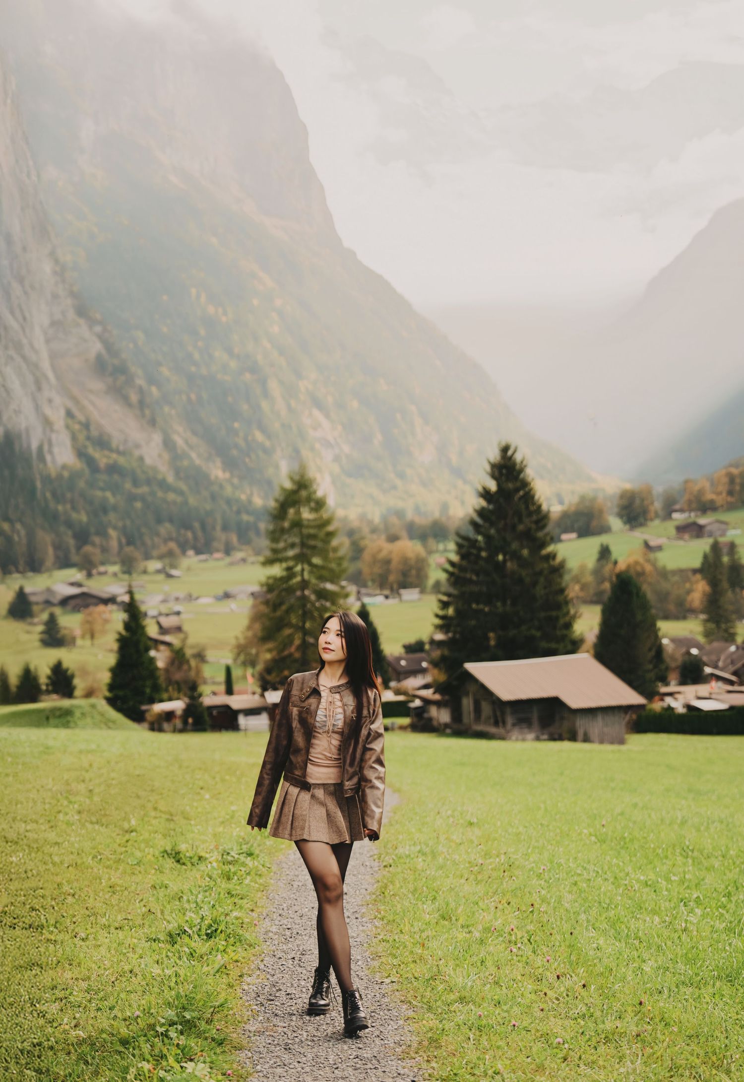 Woman in brown jacket and skirt walking on a path with a scenic Swiss mountain valley backdrop.