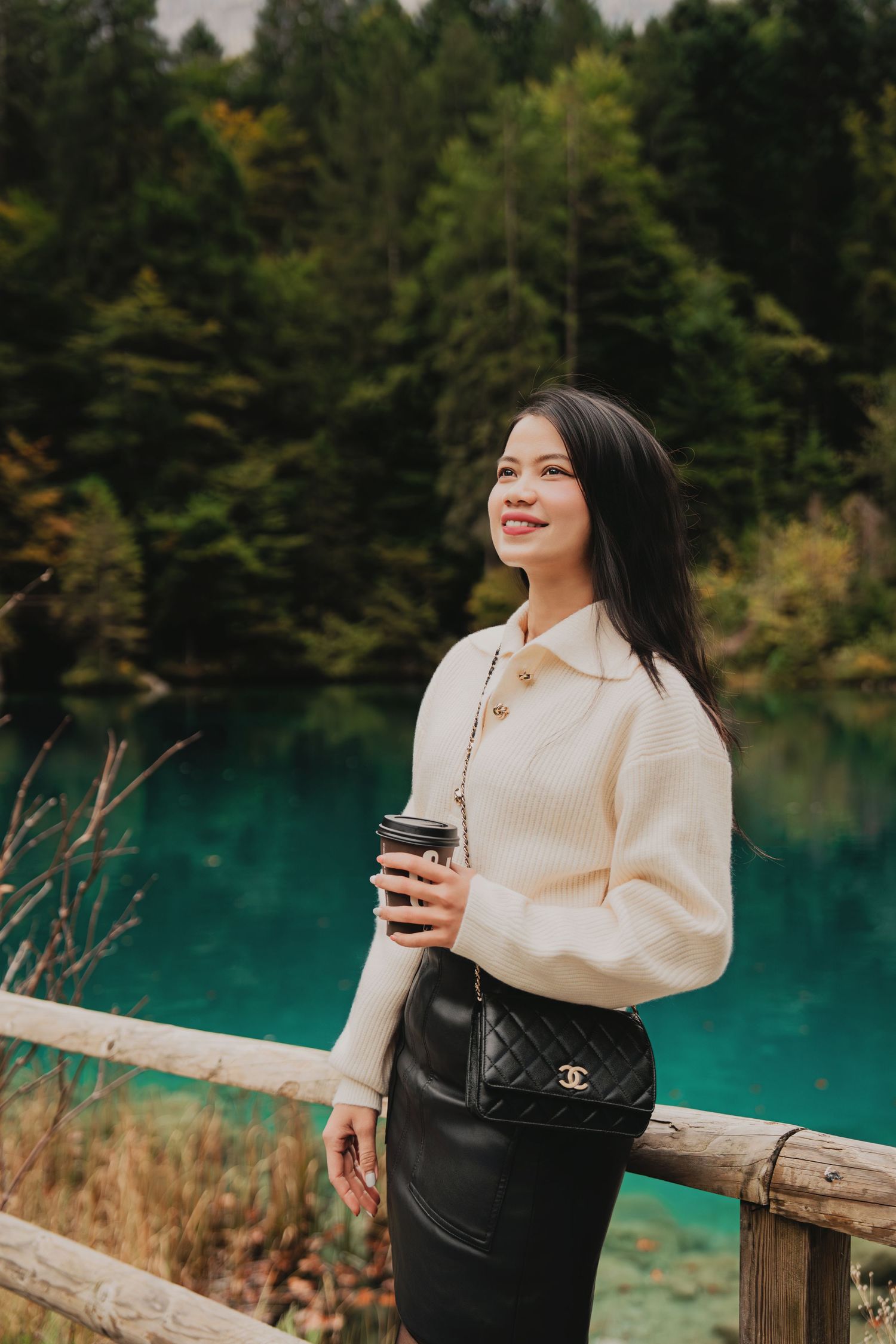 Woman in cream sweater holding coffee with Chanel bag leaning on fence by vivid turquoise alpine lake.