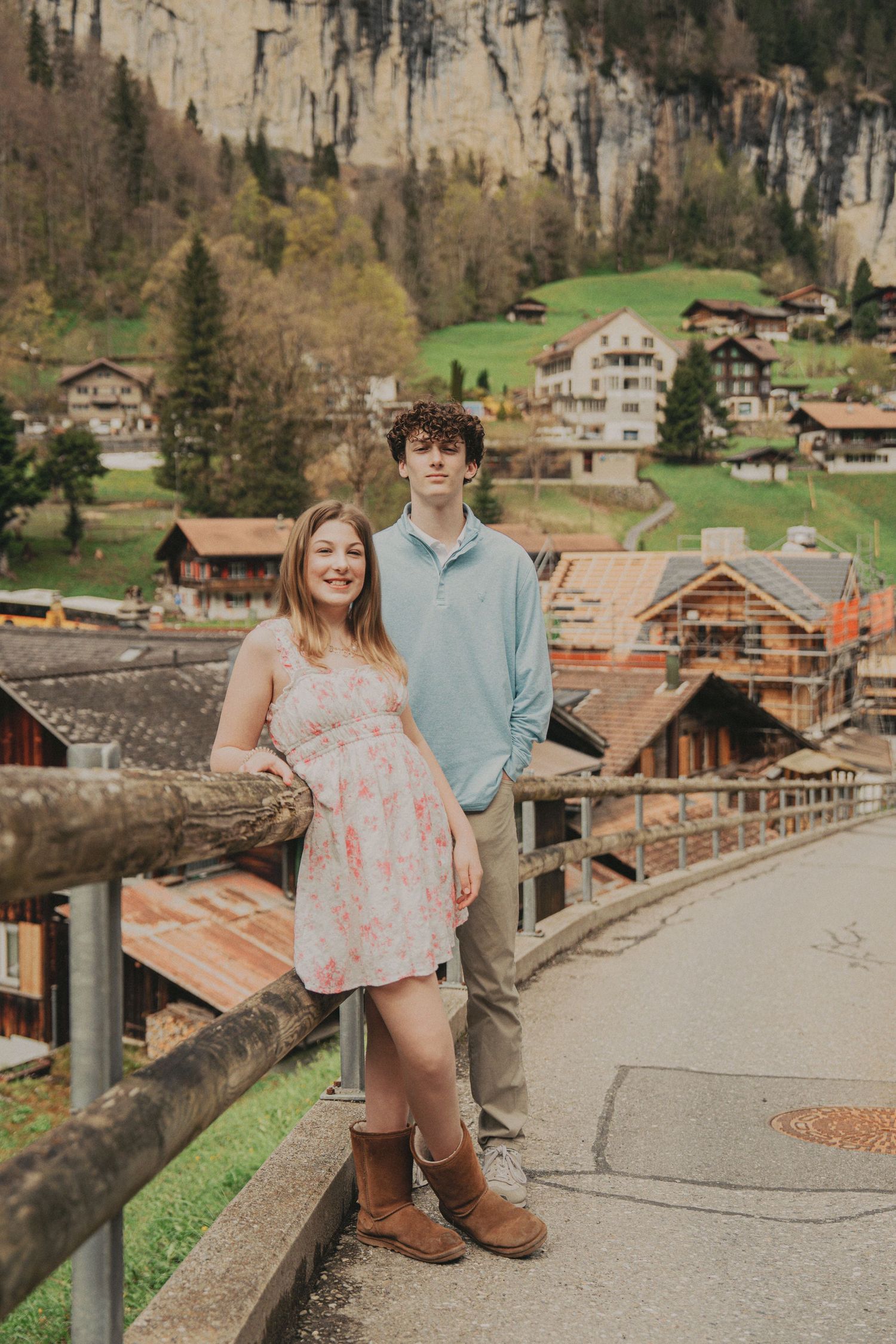 Teen boy and girl posing by a wooden railing with Swiss alpine village and cliffs behind them.