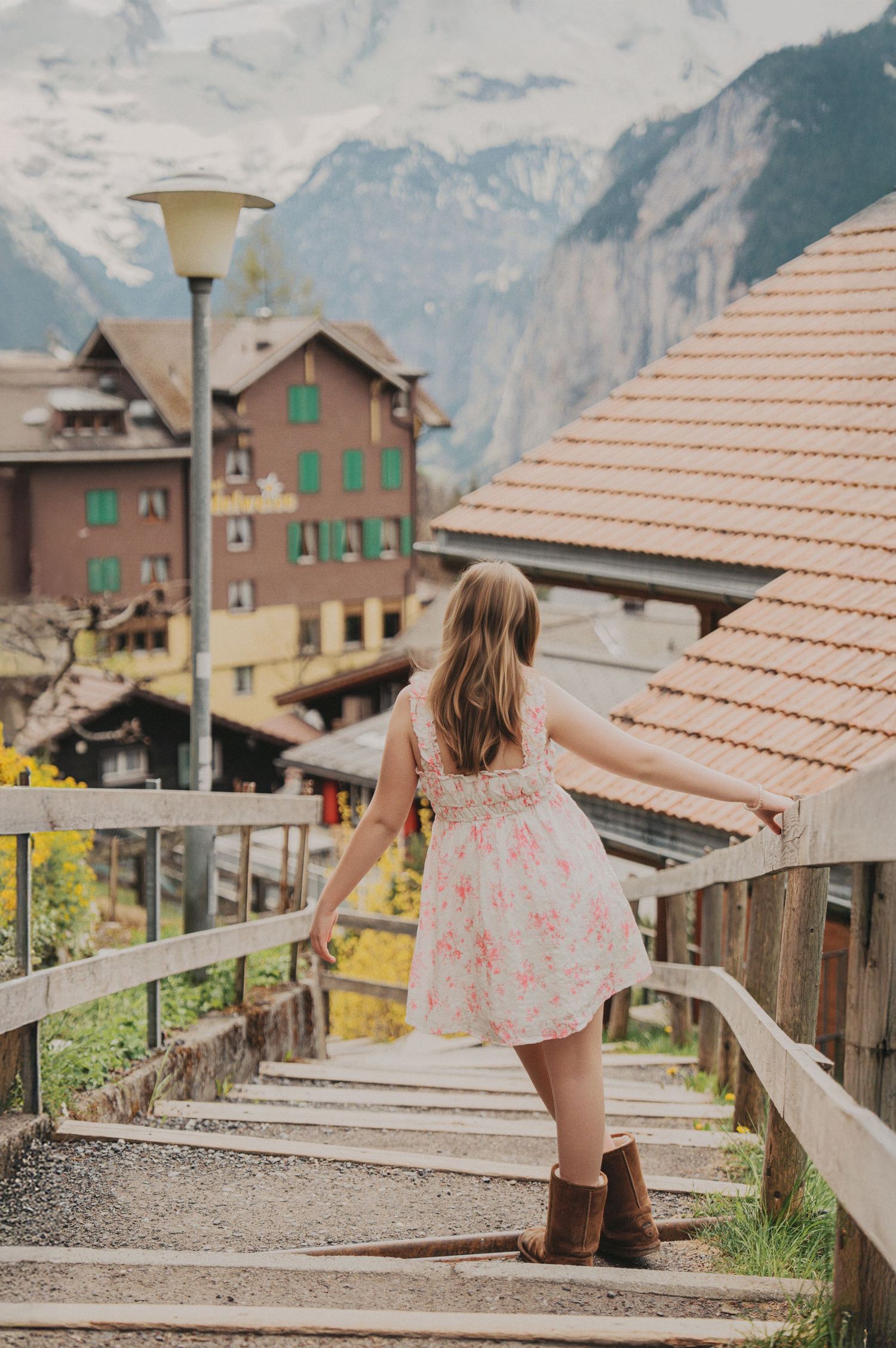 Girl in floral dress and boots walking up wooden stairs in Swiss Alpine village with mountain backdrop.