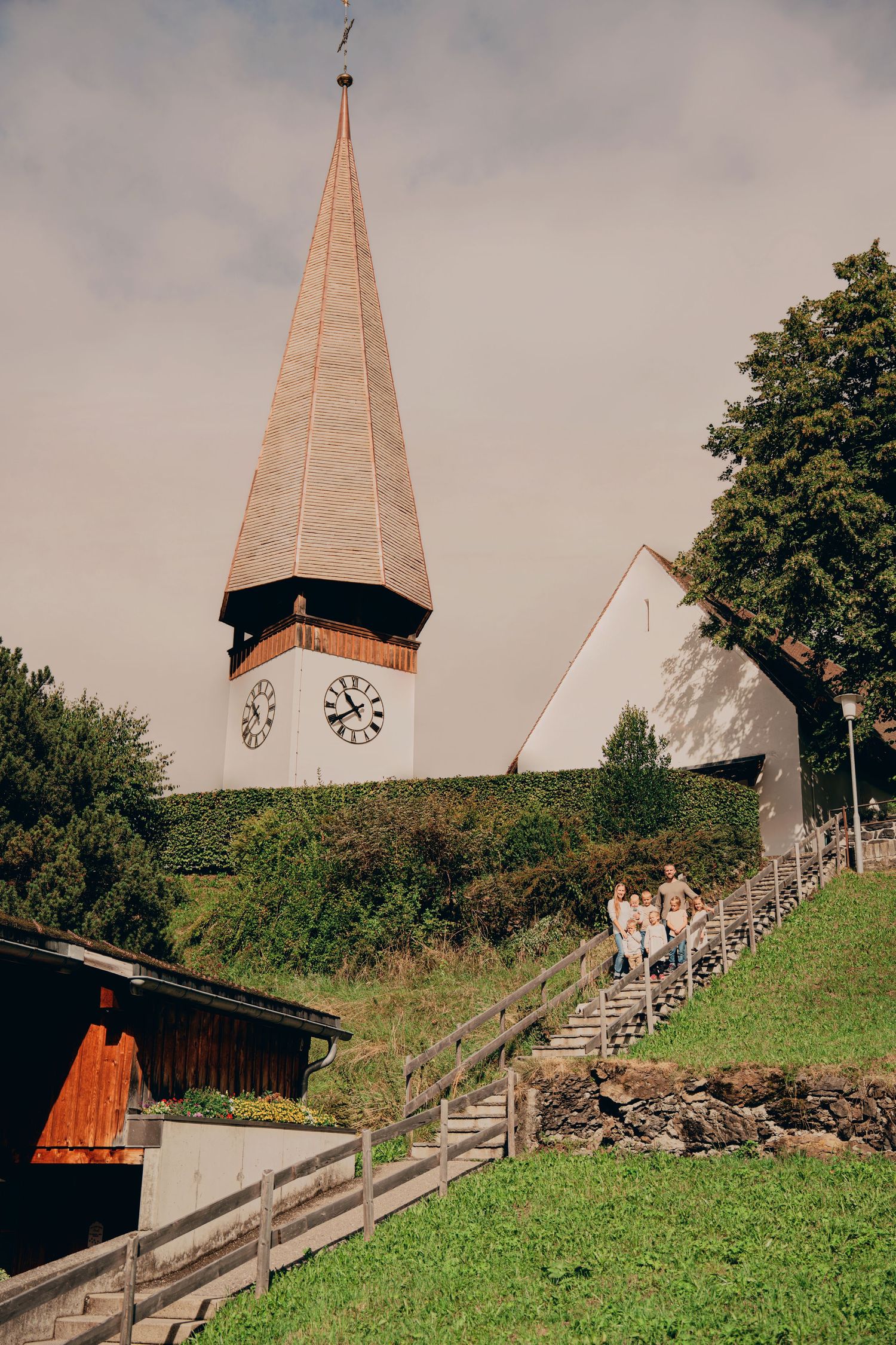 Rustic Swiss church with a pointed wooden steeple and clock tower on a hillside stairway.