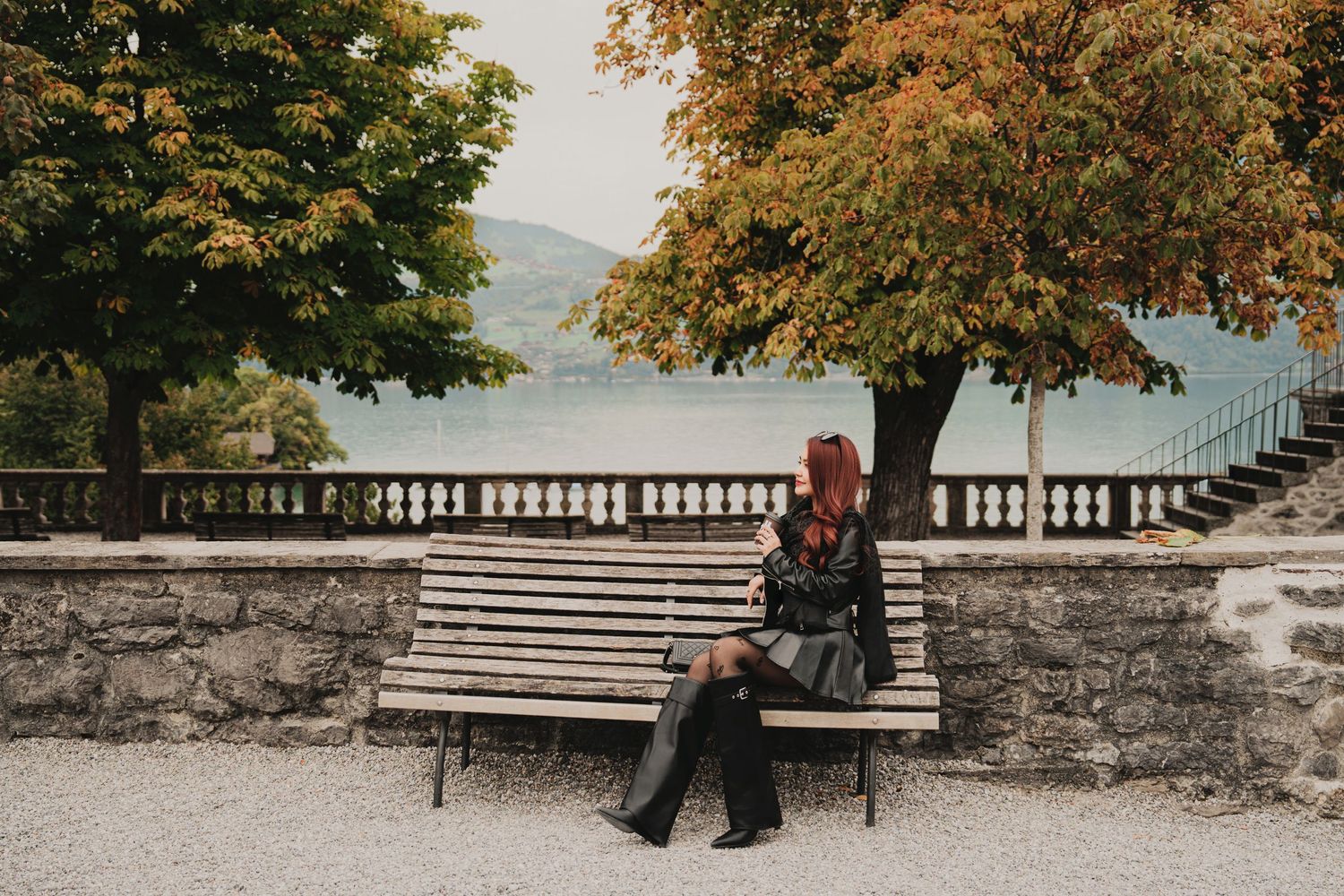 Woman with red hair sitting on park bench beside autumn trees with a tranquil lake in background.
