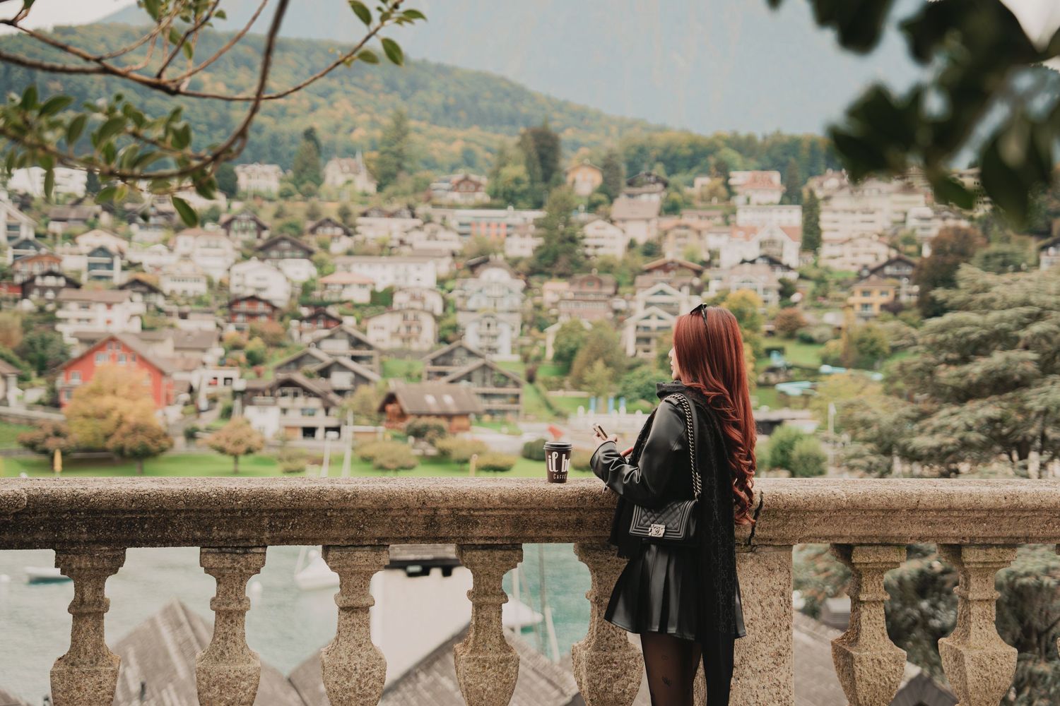 Young woman with red hair and backpack photographs picturesque lakeside Swiss town from stone balustrade.