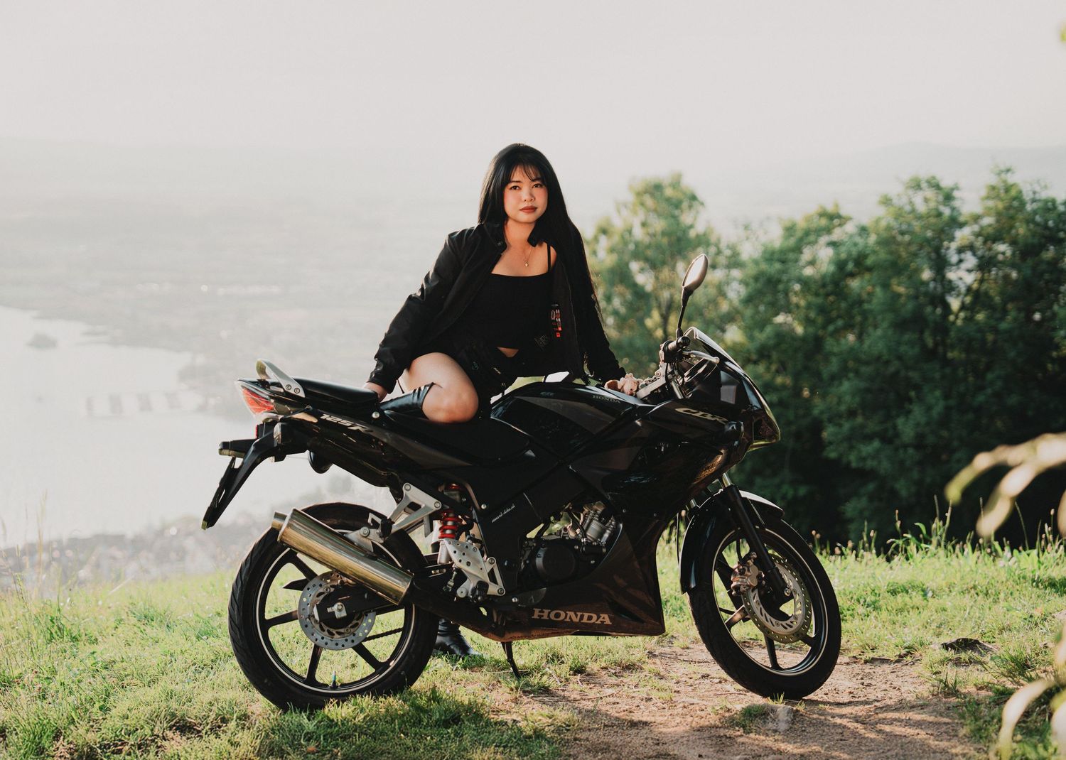 Young woman in black outfit sitting on a Honda motorcycle on a scenic hillside overlooking a river at sunset.