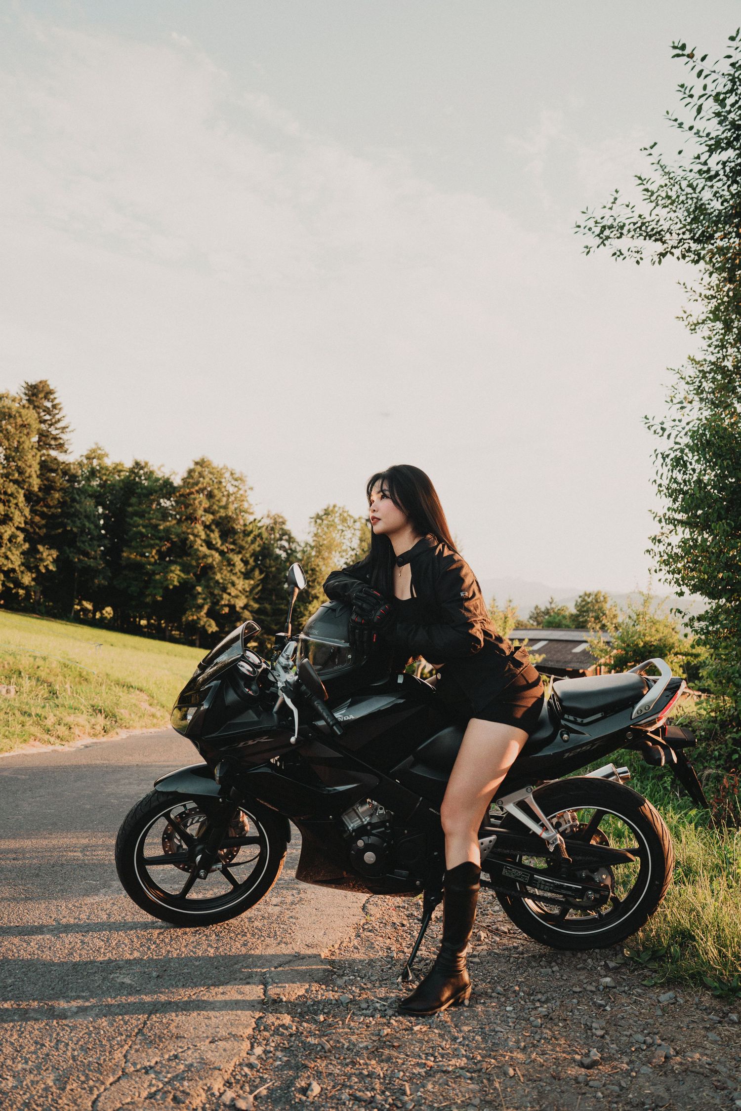 Fashionable woman in black posing with a black motorcycle on a quiet road with lush green scenery.