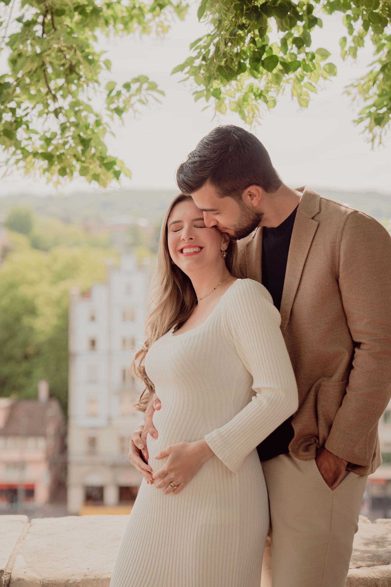 Man in tan blazer tenderly kisses pregnant partners head while she smiles with eyes closed outdoors.