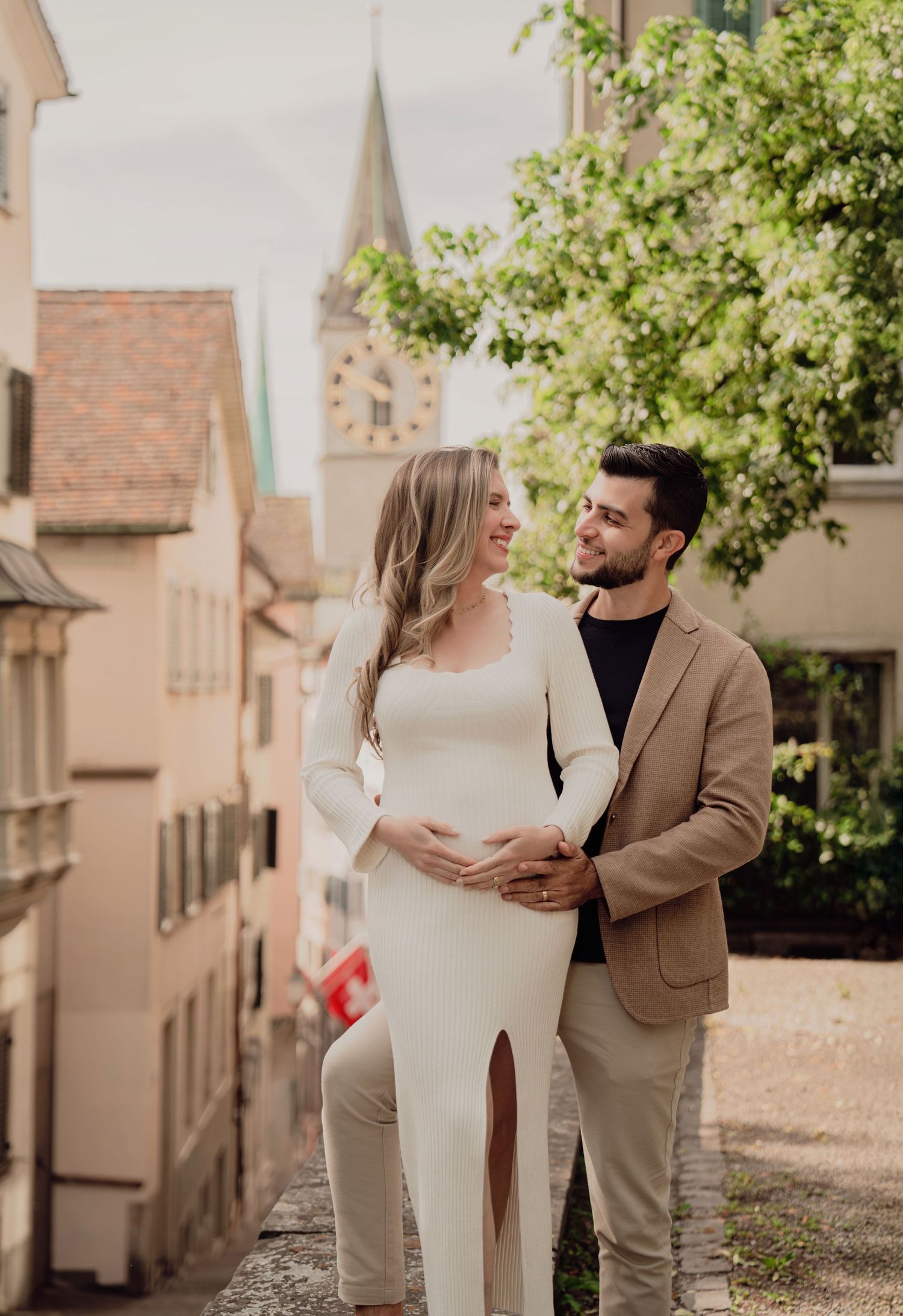 Pregnant couple embracing on a European cobblestone street with a church steeple in the background.