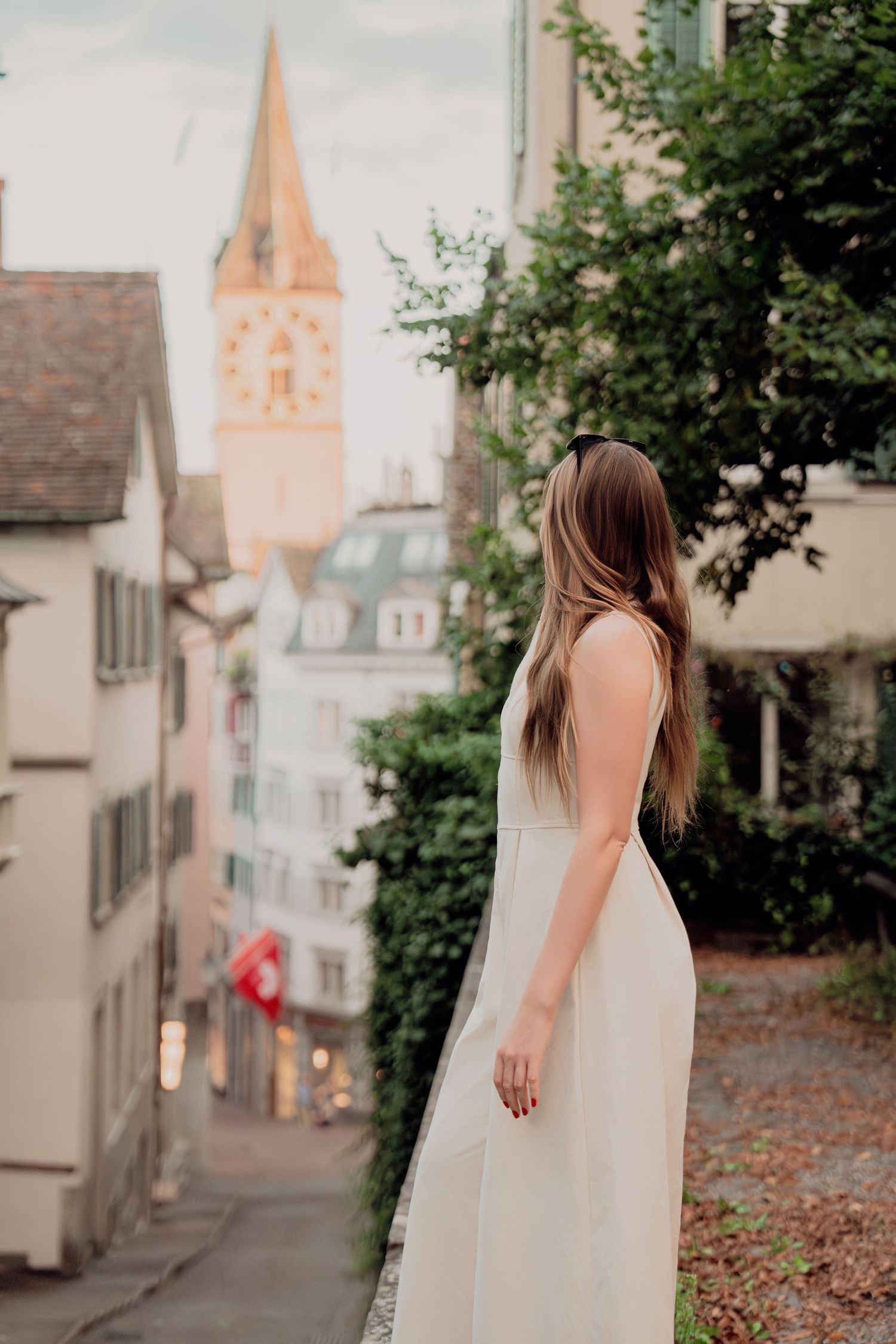 Woman in white dress viewed from behind on charming European alley with clock tower and Swiss flag visible.