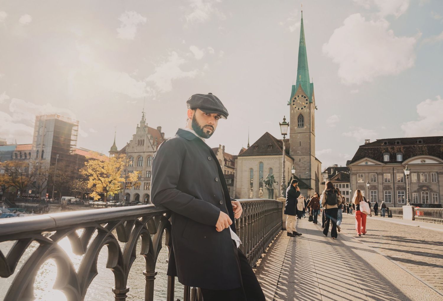 Stylish bearded man in dark coat and flat cap leaning on bridge railing with Zurich church spire behind.