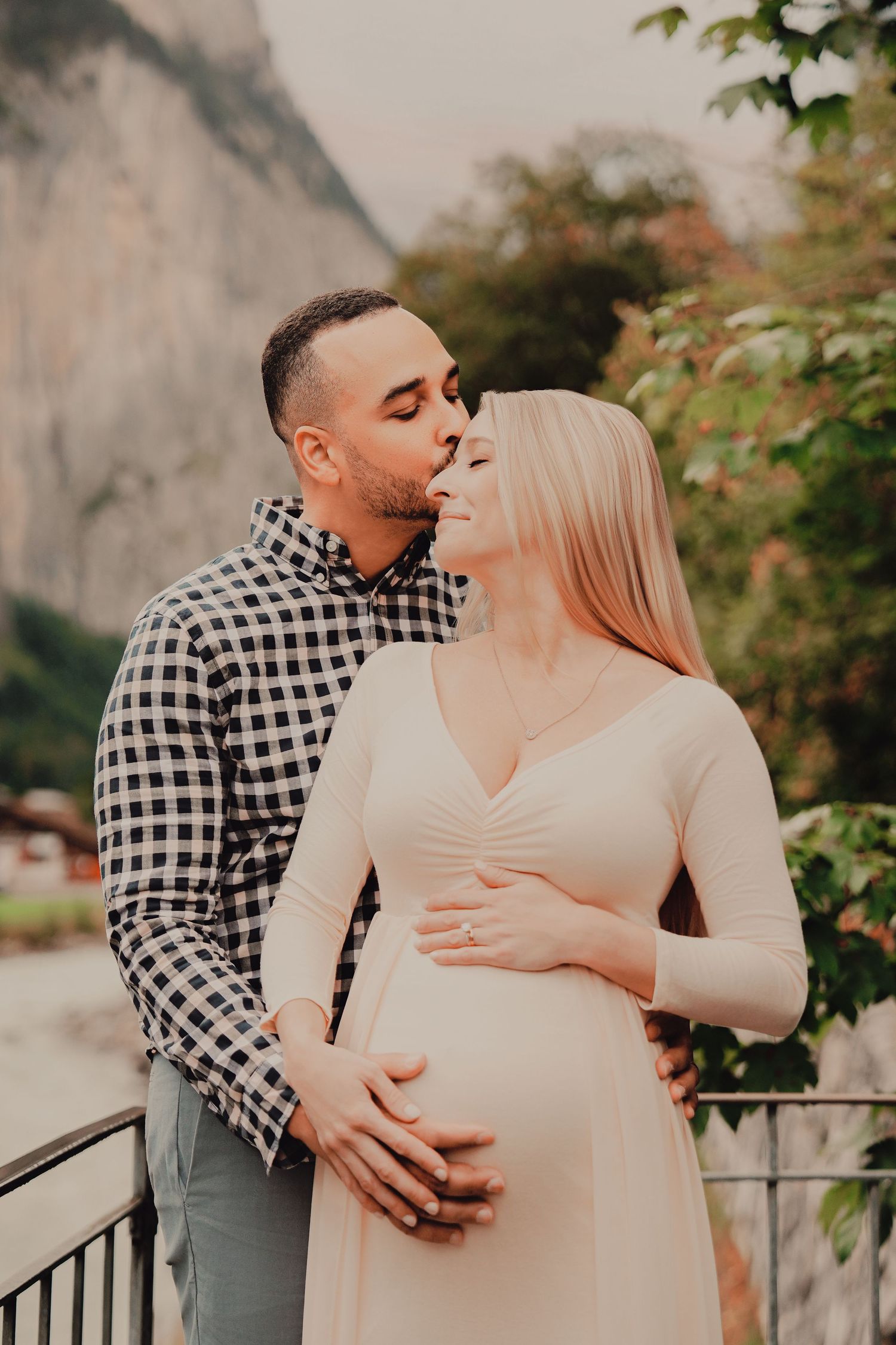 Man kissing pregnant woman's head while holding her belly on balcony with mountain backdrop.