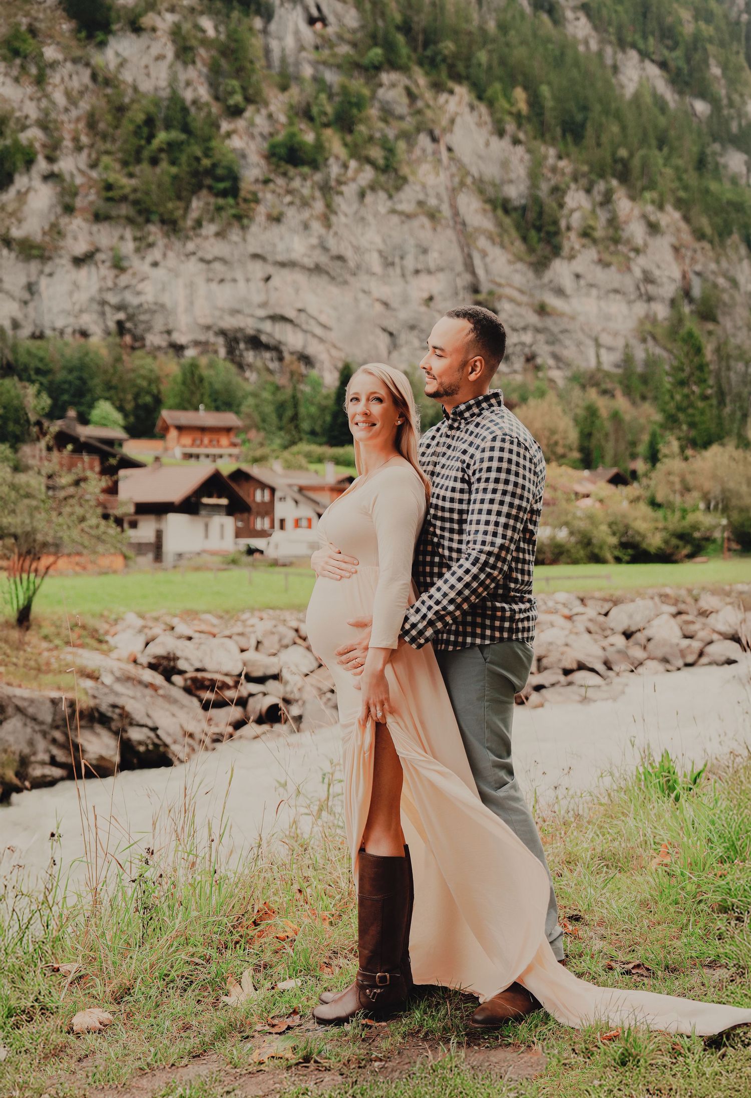 Smiling pregnant woman in blush gown embraced by partner during maternity photoshoot near rocky Swiss mountain stream.