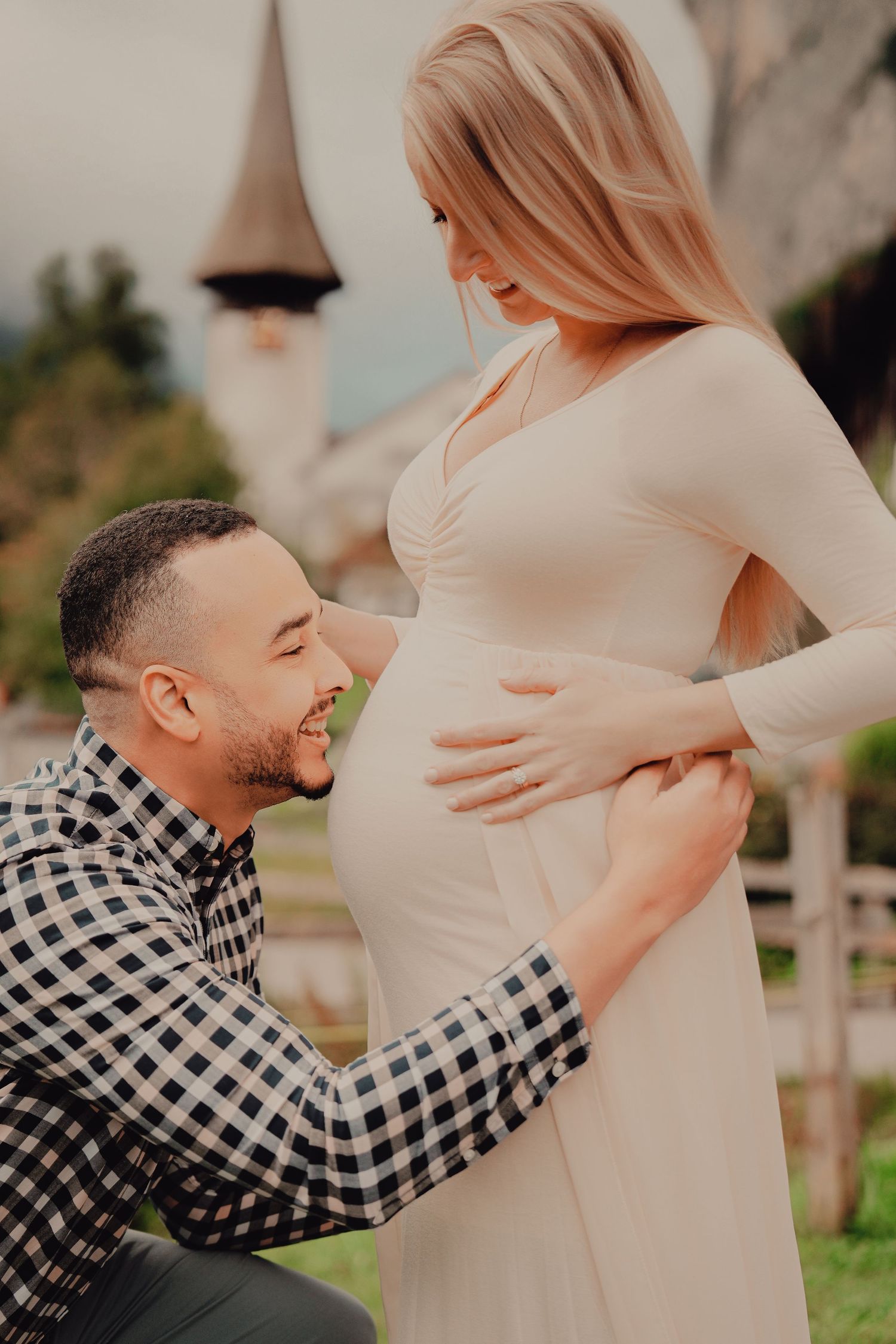 Man kneels smiling at pregnant womans belly while she cradles it outdoors in a Swiss mountain setting.