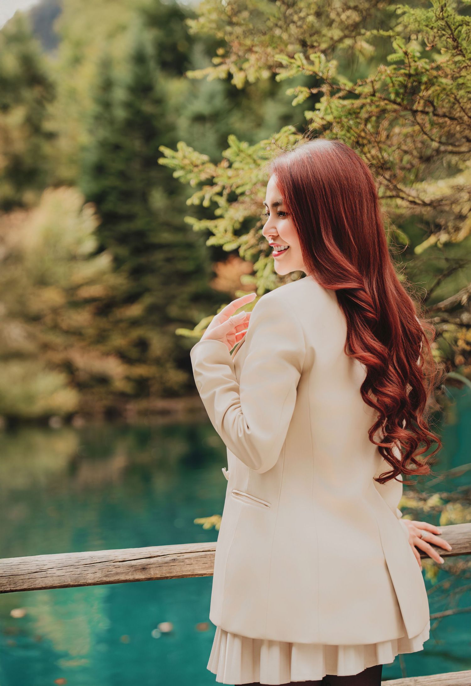 Smiling red-haired woman in cream blazer posing by turquoise lake with autumn forest backdrop.