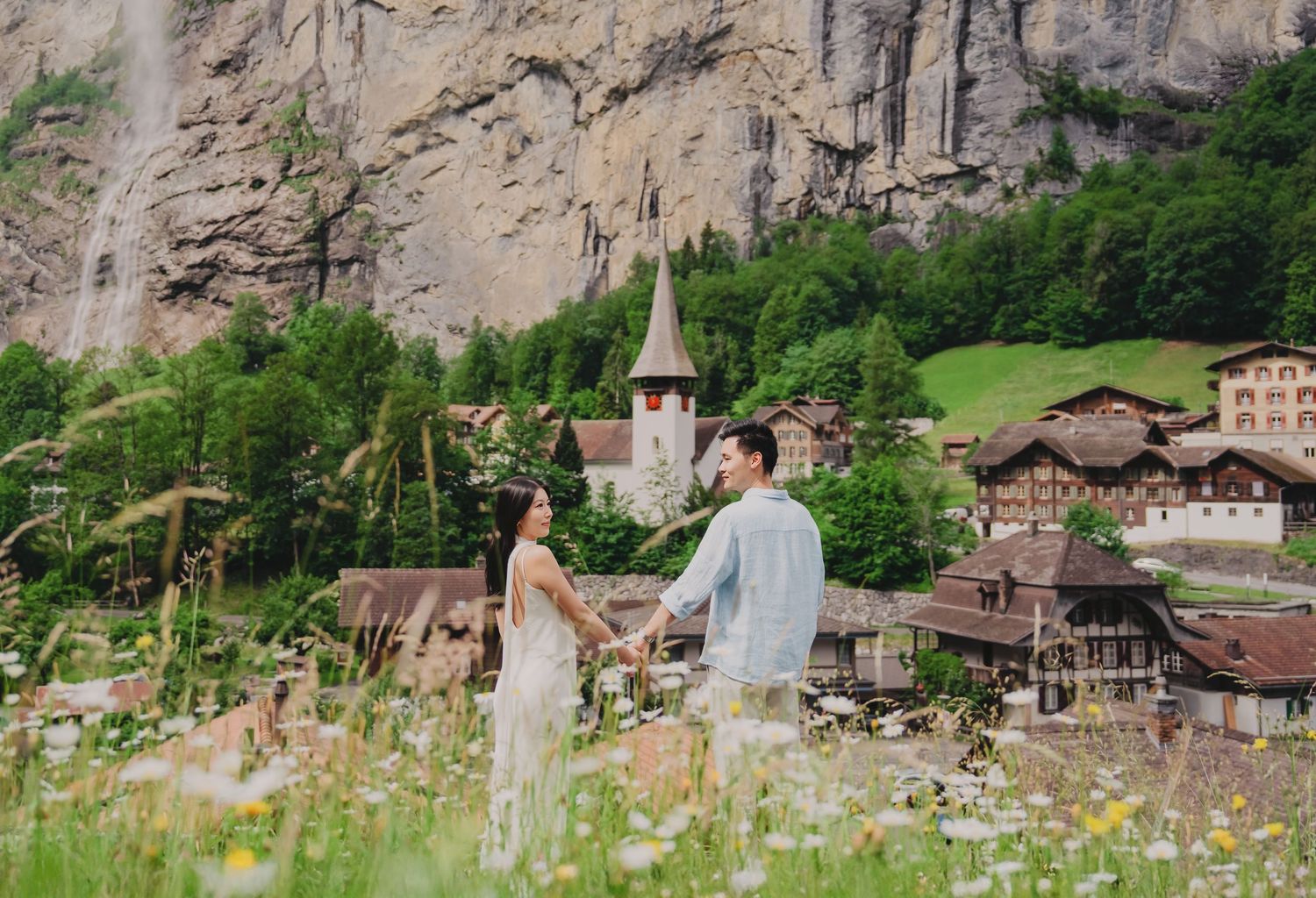 Couple holding hands in a wildflower meadow with Swiss village church and waterfall backdrop in Lauterbrunnen.