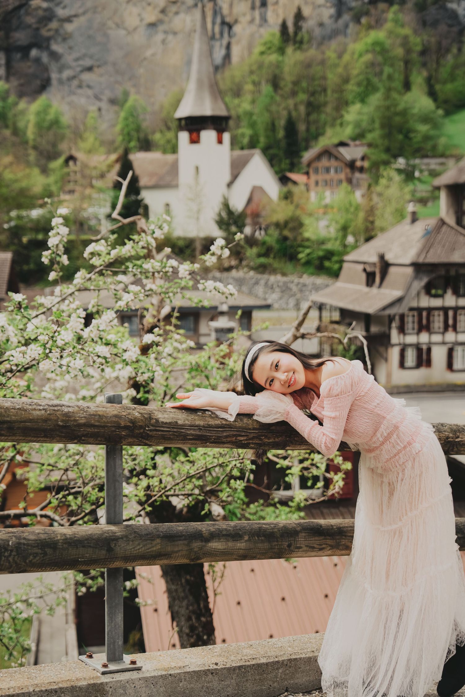 Smiling woman in pink dress resting on wooden fence with Lauterbrunnen church and blooming trees behind.