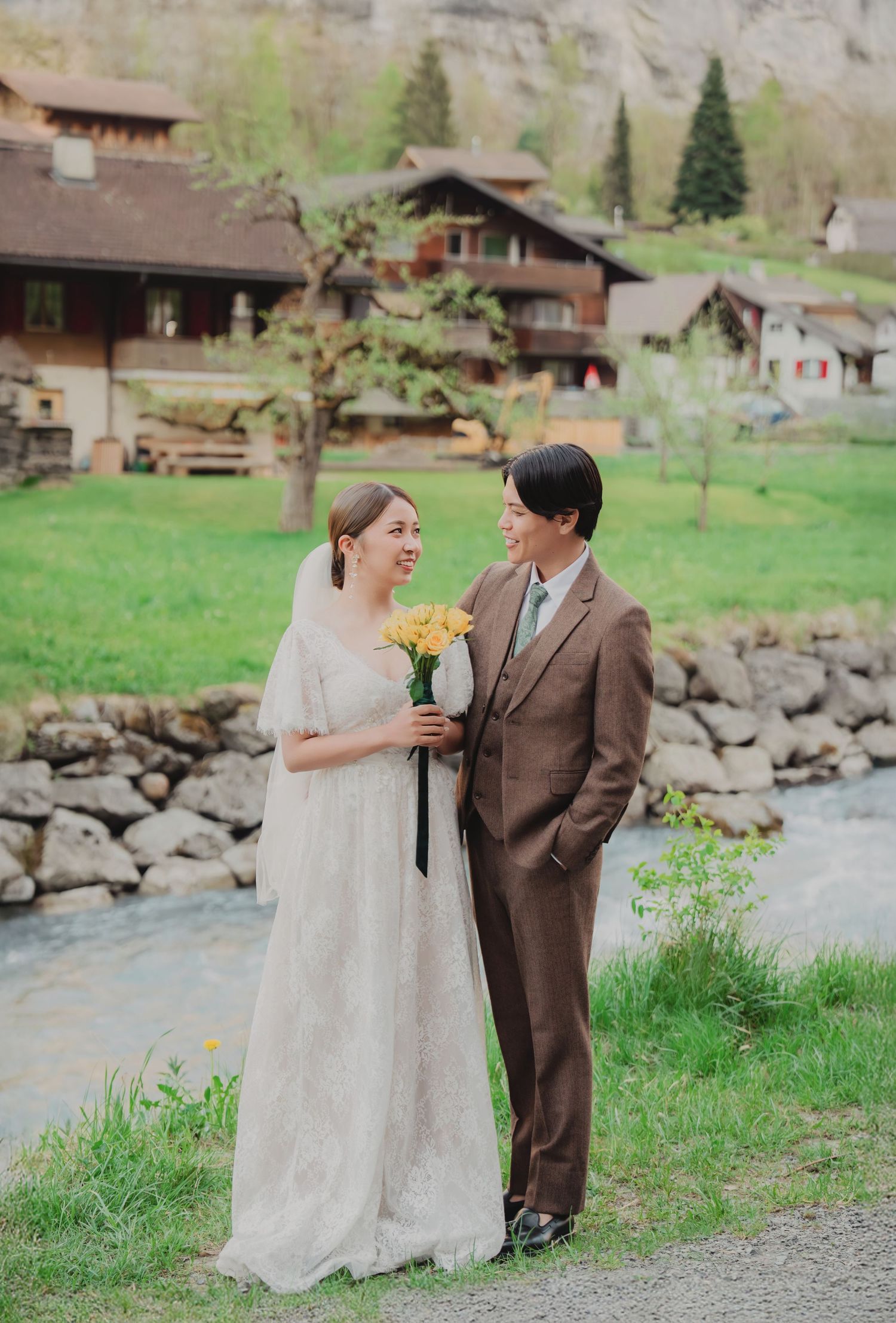 Smiling bride holding yellow bouquet and groom in brown suit stand together by a rocky riverside in Switzerland.