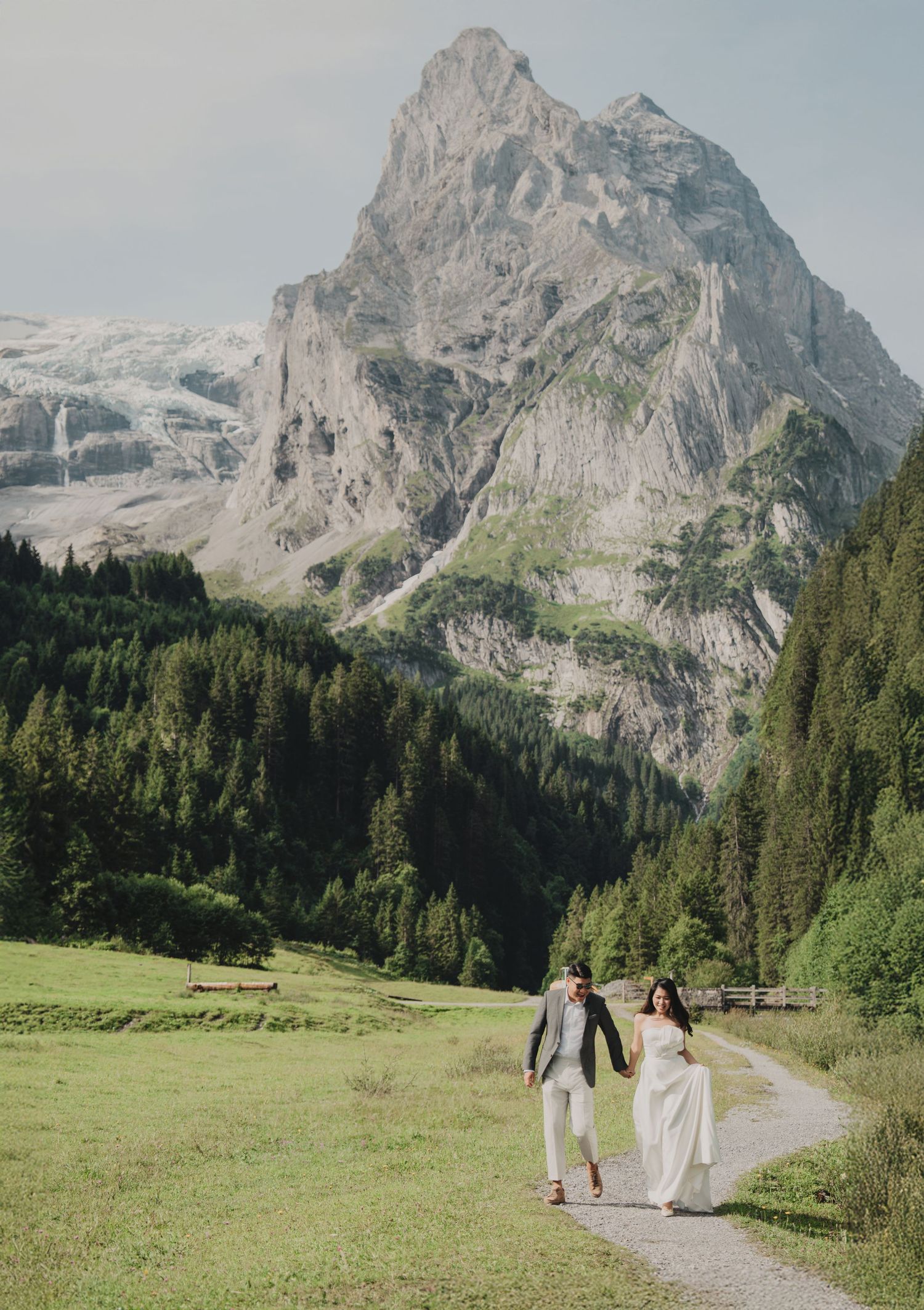 Wedding couple walking hand in hand on a mountain path with a dramatic rocky peak and glacier backdrop.