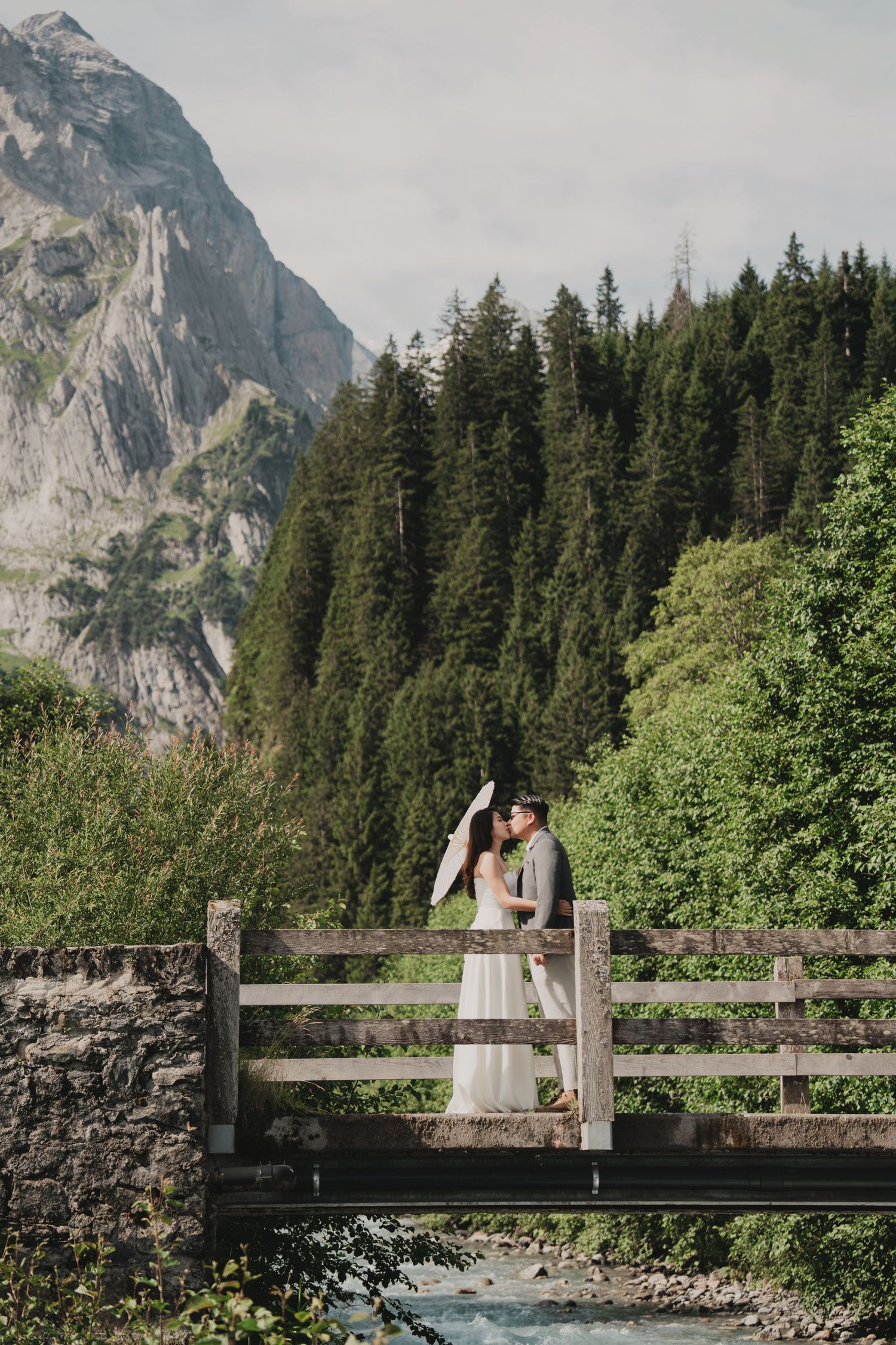 Bride and groom sharing a kiss on a rustic wooden bridge over a mountain stream with white parasol.