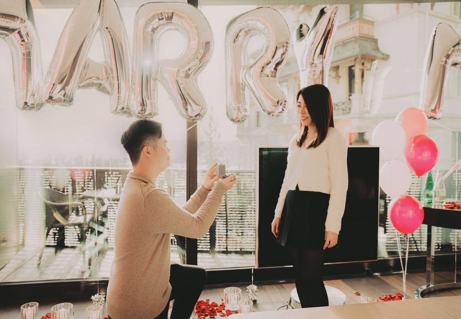 Man kneeling proposing to woman with MARRY silver balloons and pink balloons in romantic setting.