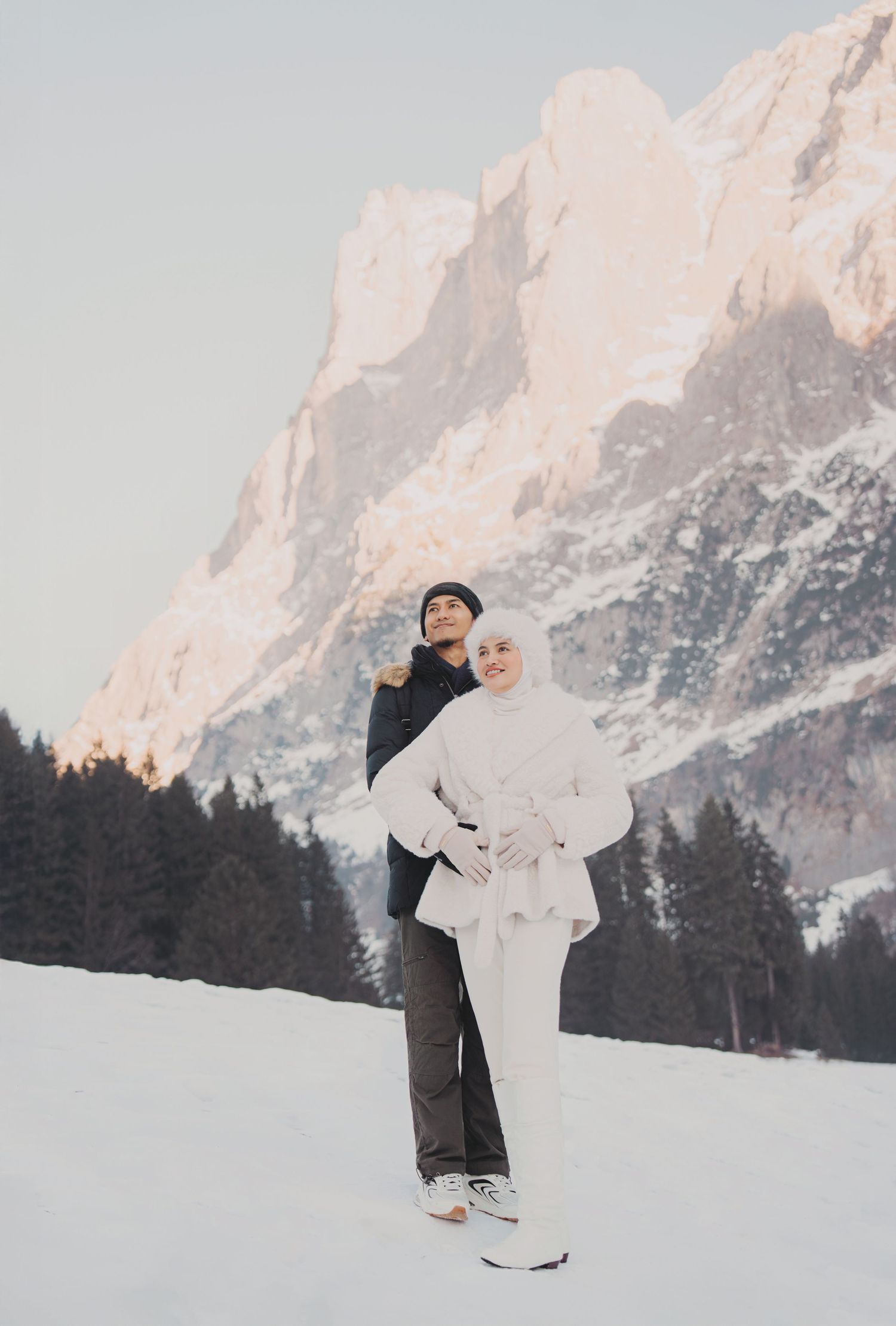 Man lifting woman in white hijab outfit against golden snow-capped mountain and pine forest backdrop.