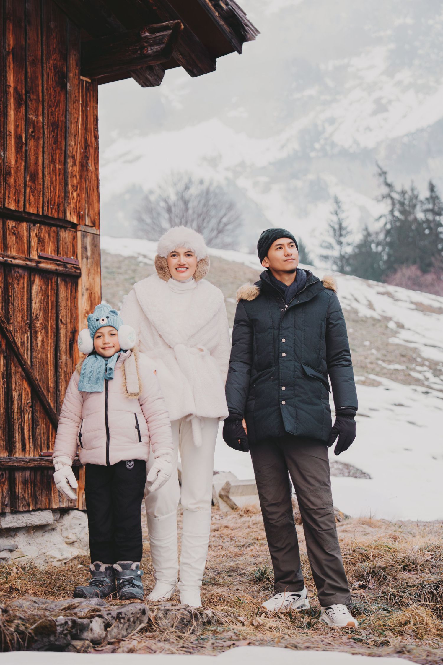 Family of three in winter coats posing beside rustic wooden alpine barn with snowy mountains.