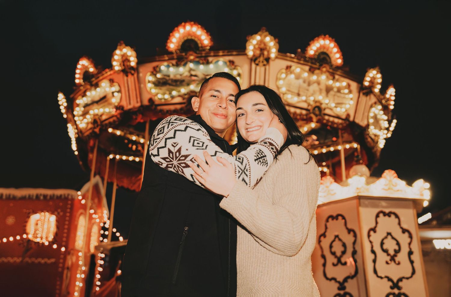 Couple hugging romantically in front of a beautifully illuminated vintage carousel at a Christmas fair.