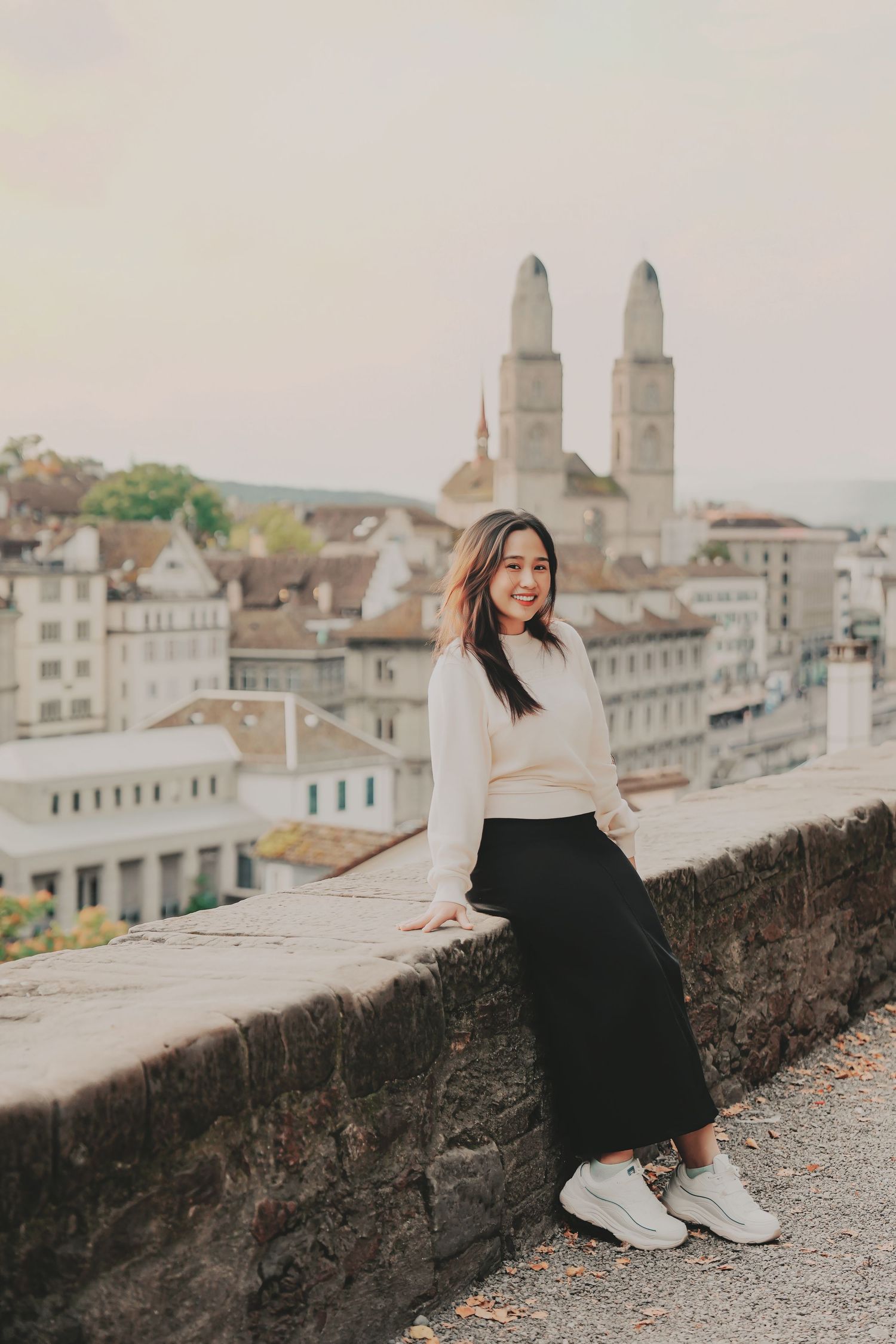 Young Asian woman in cream sweater and black skirt leaning on stone wall with Zurich skyline.