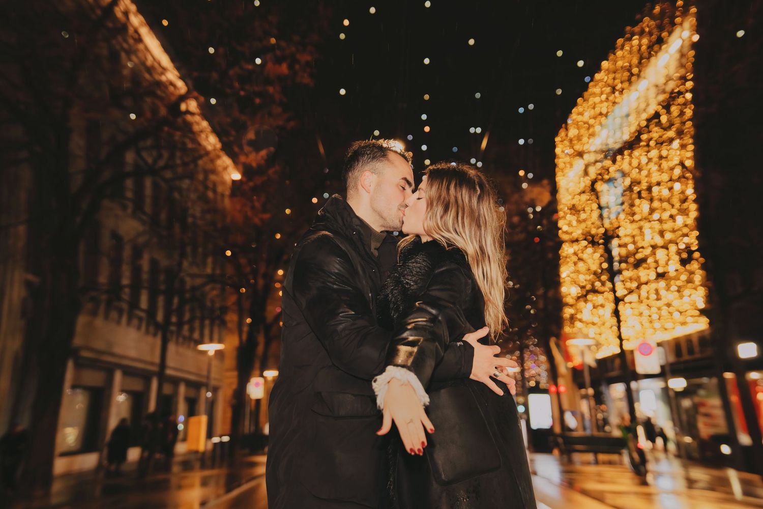 Couple kissing under sparkling Christmas lights on a rainy city street at night.
