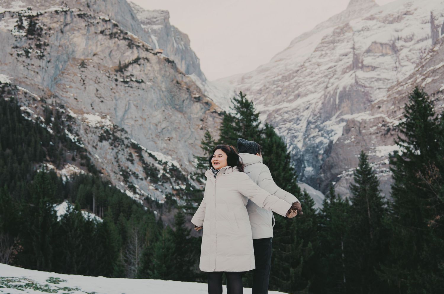 Woman in white puffer coat standing with arms outstretched among snow-covered trees and mountains.