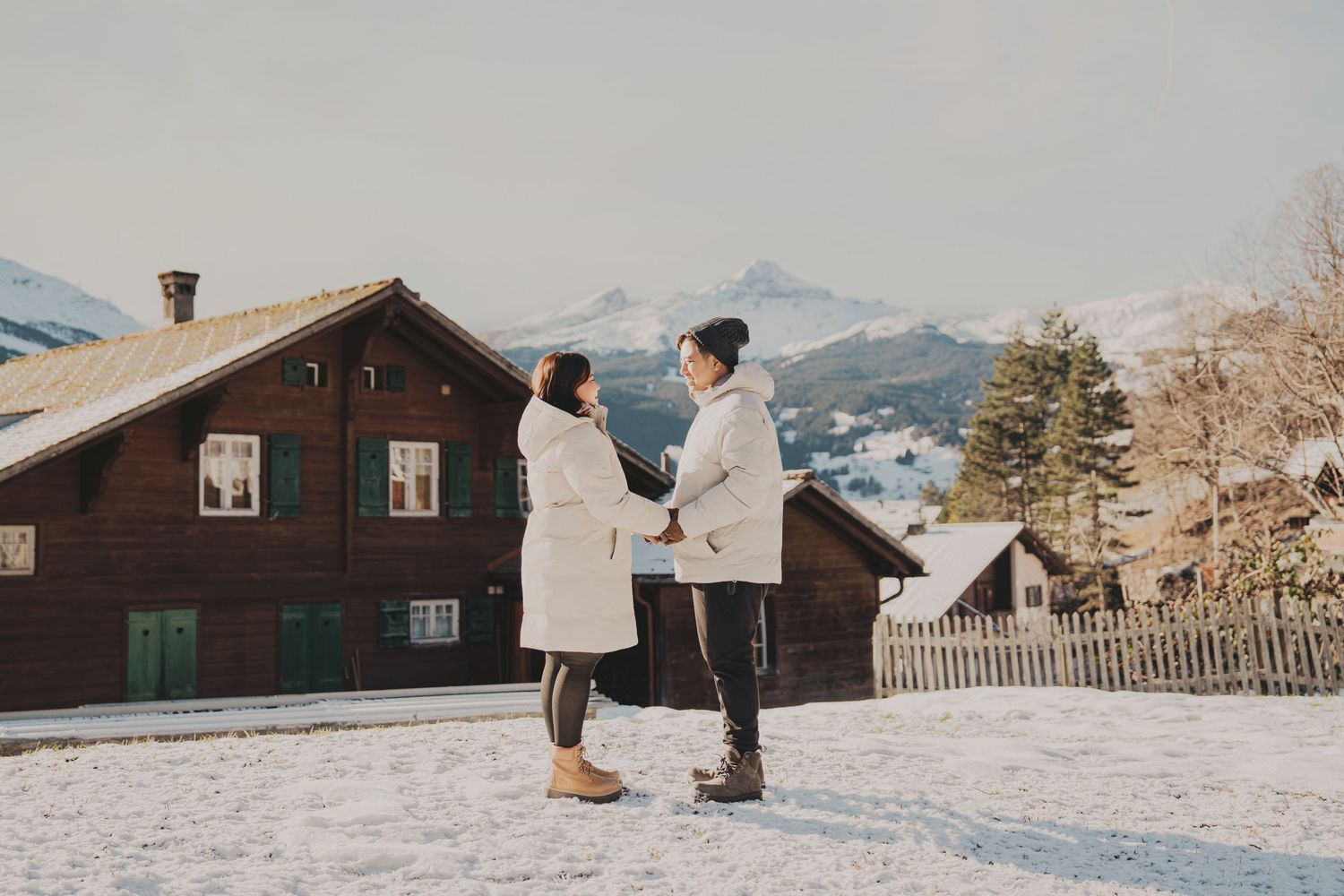 Couple in white coats holding hands outside a rustic brown Swiss chalet surrounded by winter snow.